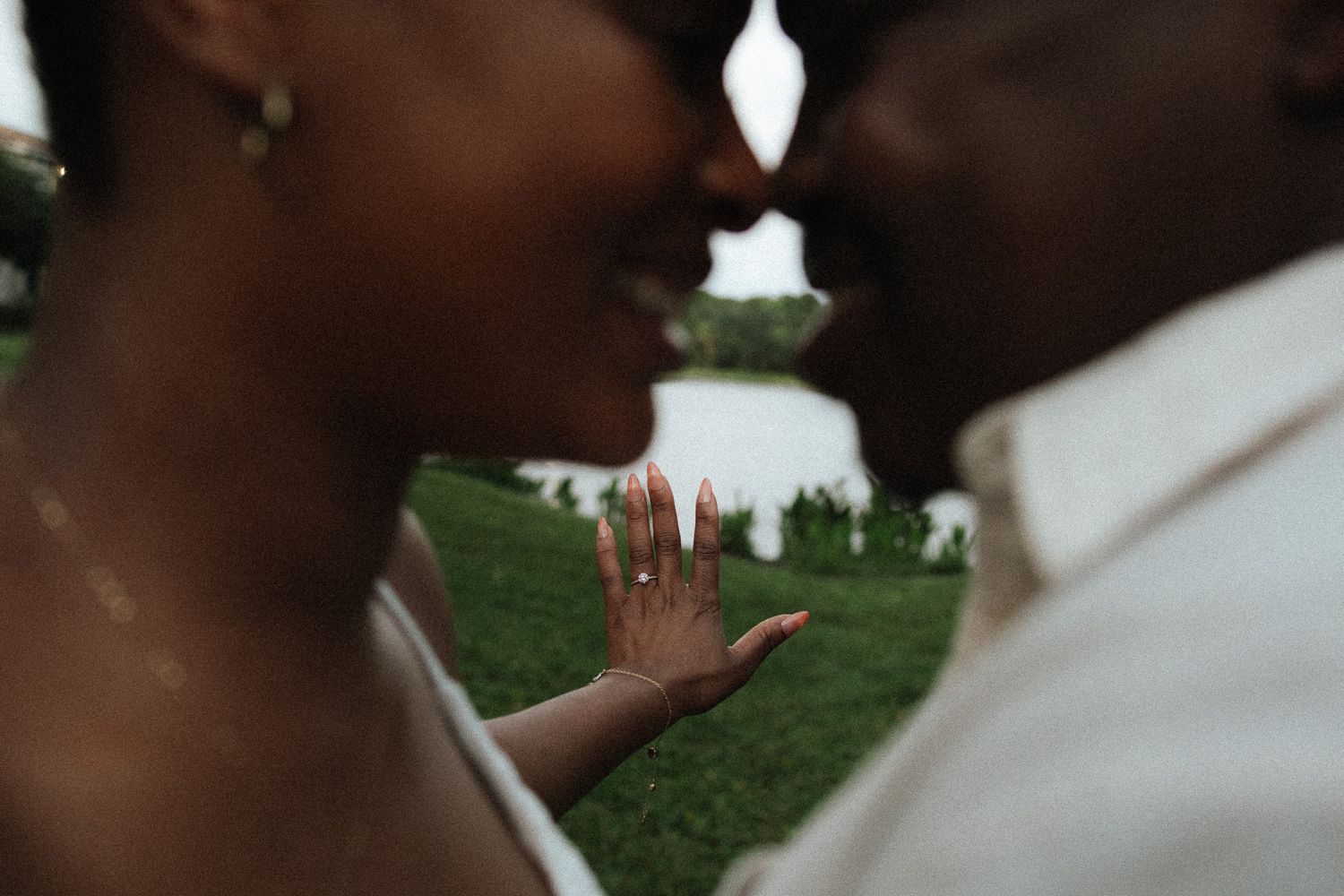 An engaged couple smiles closely together as the woman displays her sparkling diamond ring near a lake.