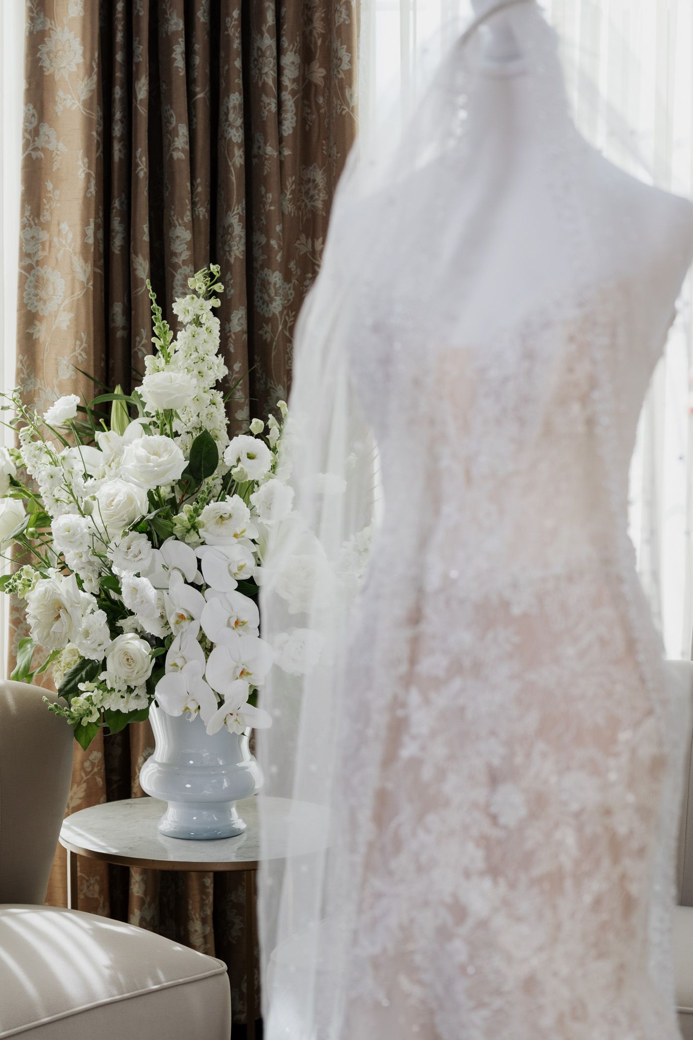 Lace bridal gown on mannequin beside a vase of white orchids and flowers in a suite.