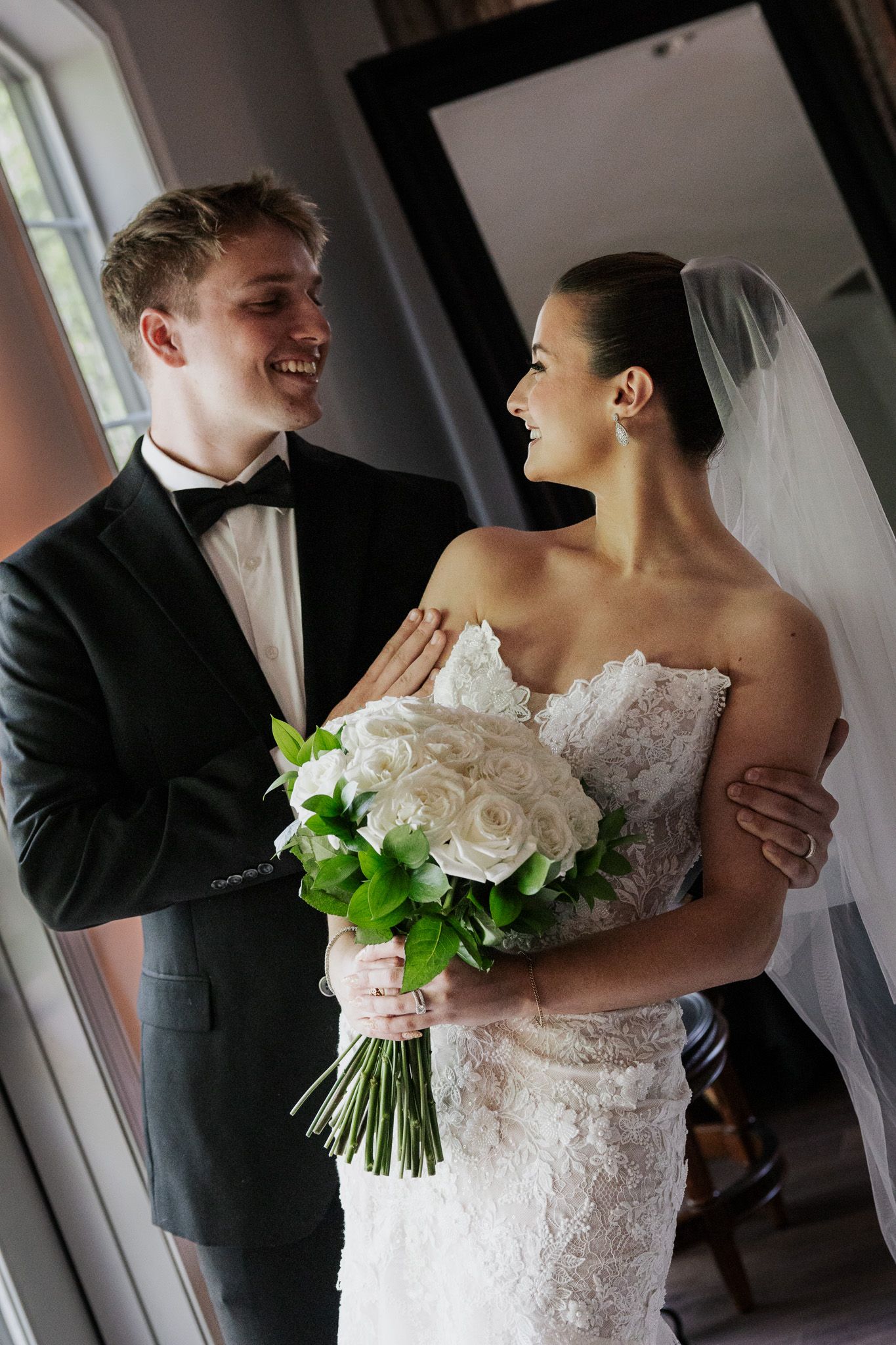 Joyful bride and groom laugh together, bride holding a white rose bouquet in a lace gown with veil.