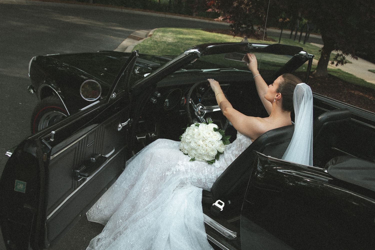 Bride in lace gown seated in vintage black convertible holding white bouquet on wedding day.