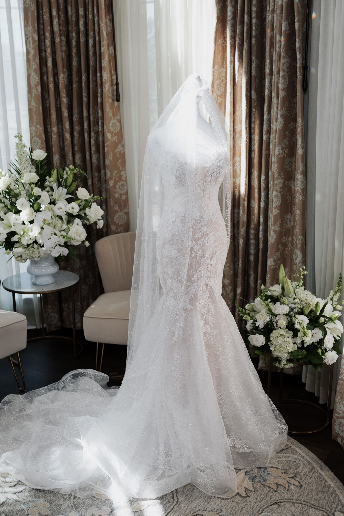 Elegant lace mermaid wedding gown with cathedral veil on mannequin flanked by white floral arrangements.