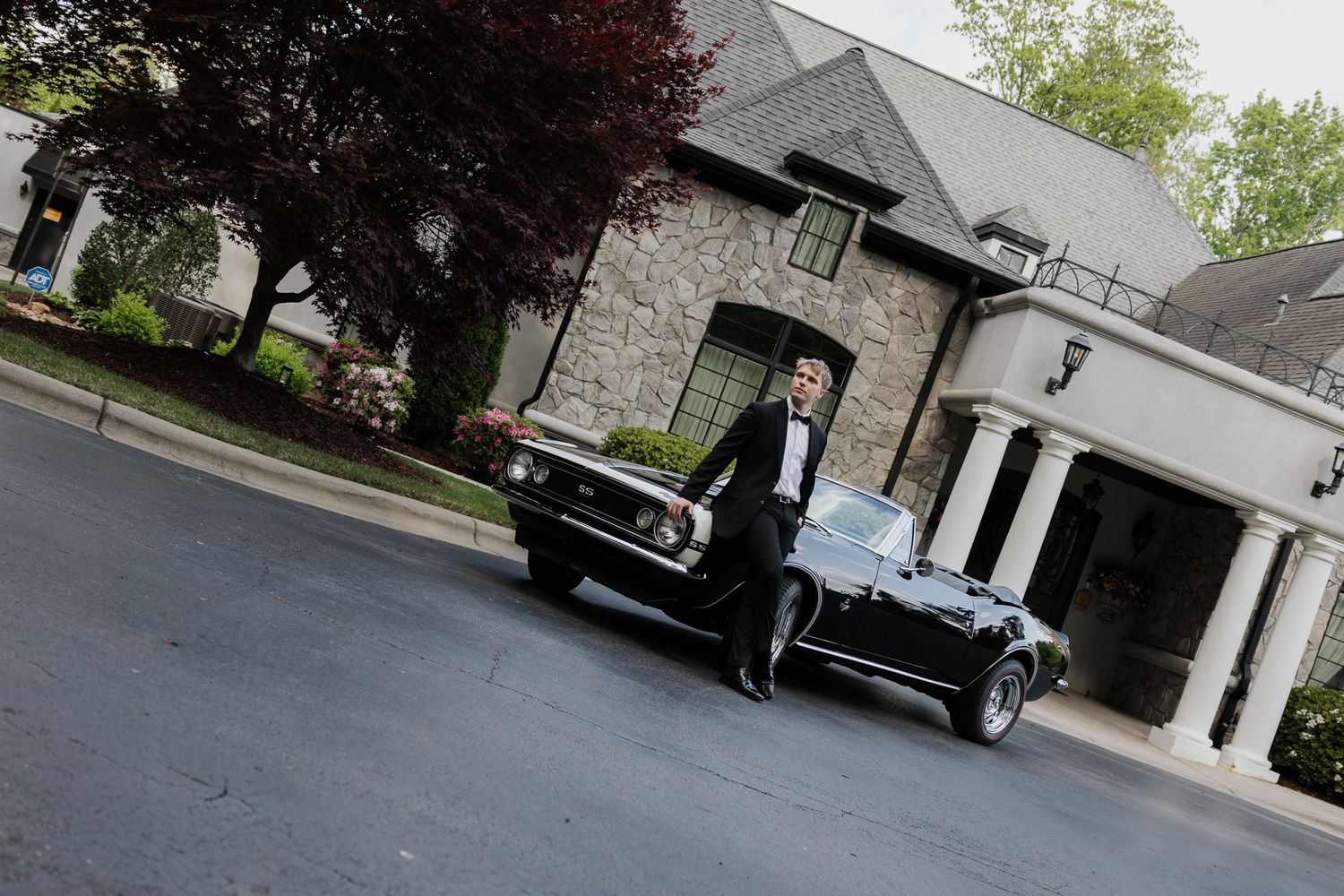 Groom in tuxedo standing beside a classic black convertible in front of a stone estate.