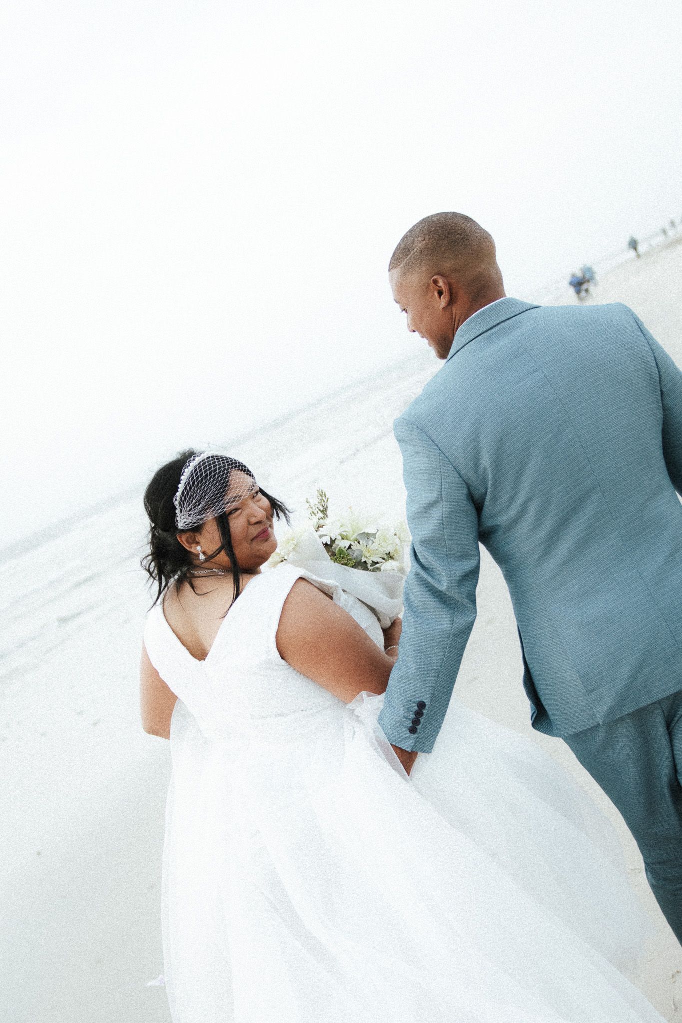 Bride and groom walking hand-in-hand on a misty beach, bride holding a white bouquet.