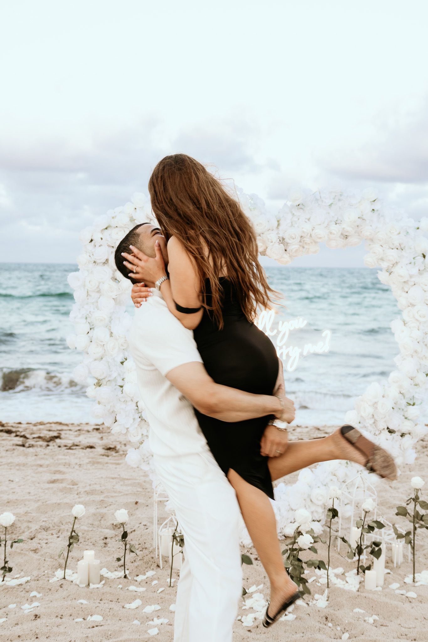 Man lifting woman in black dress during beach proposal with white floral arch and candles.