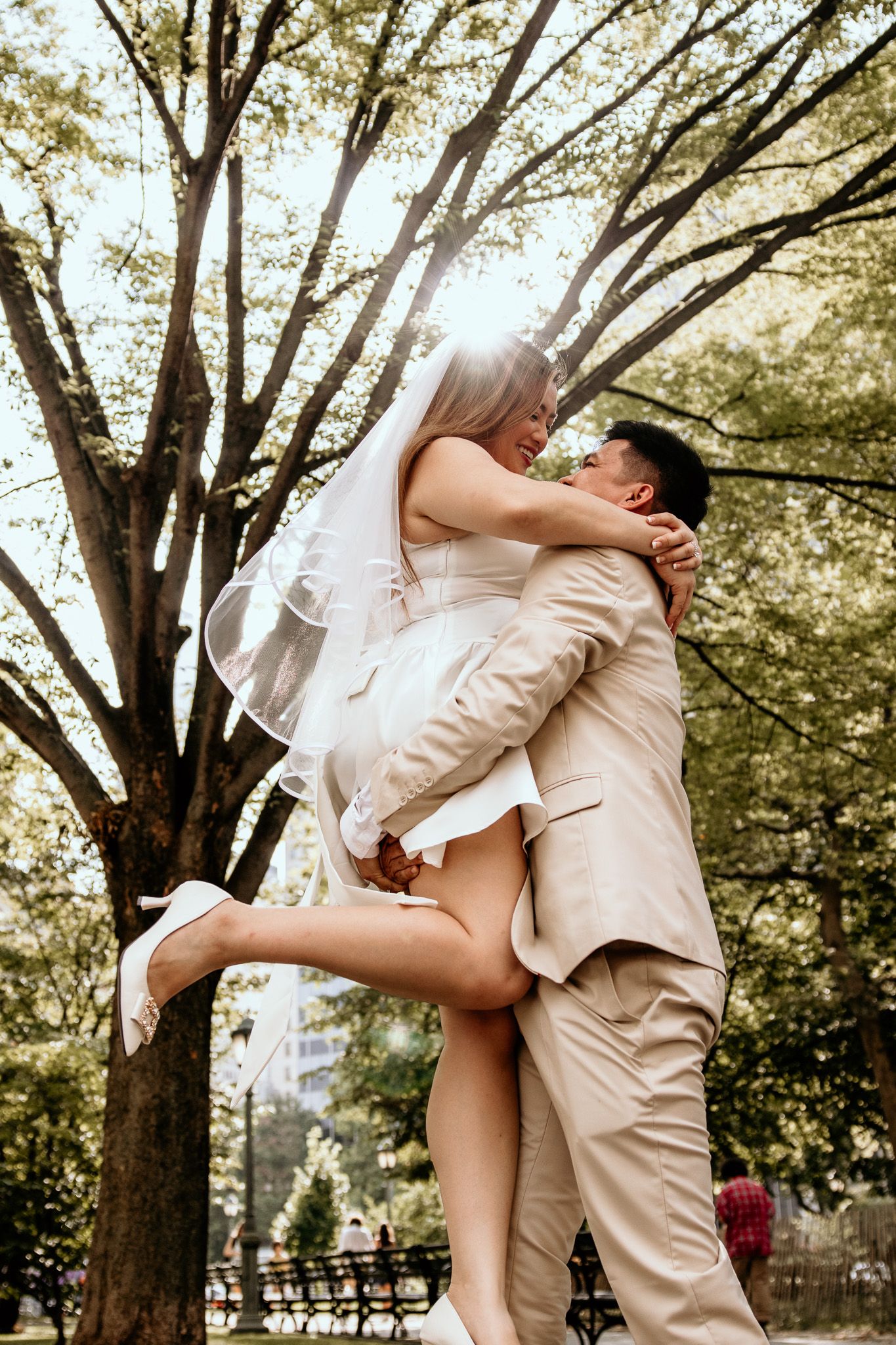 Groom in beige suit lifts bride in white mini dress and veil, sharing a joyful moment in a sunlit park.
