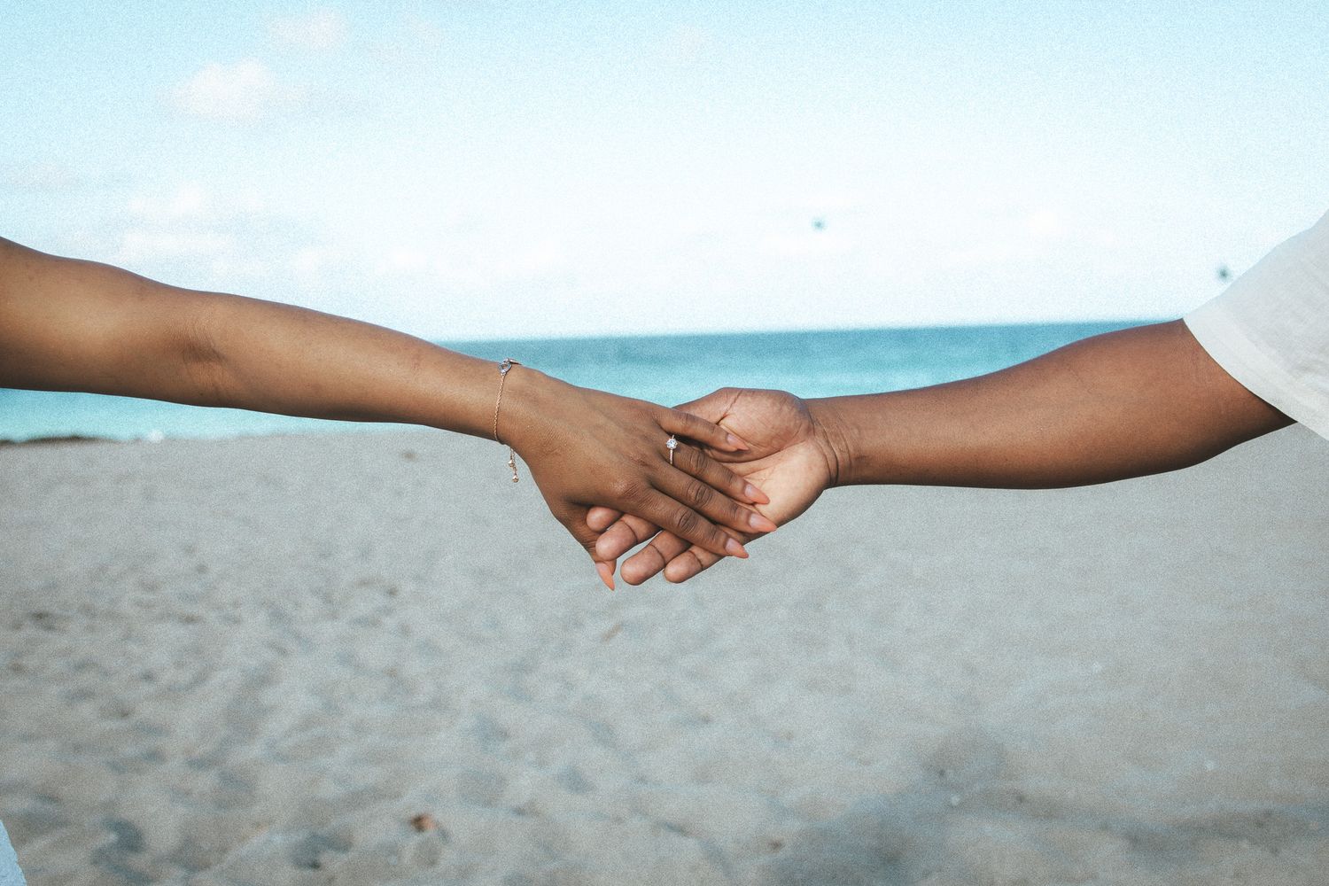 Two people holding hands on a sandy beach, with an engagement ring visible on the woman's finger.