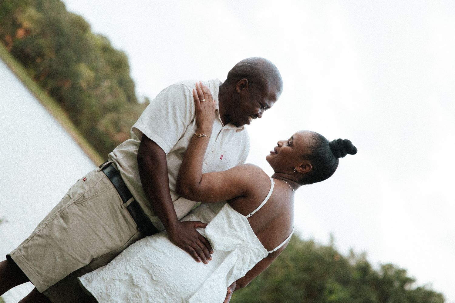A couple sharing a romantic near-kiss embrace outdoors near a lake, both dressed in white.