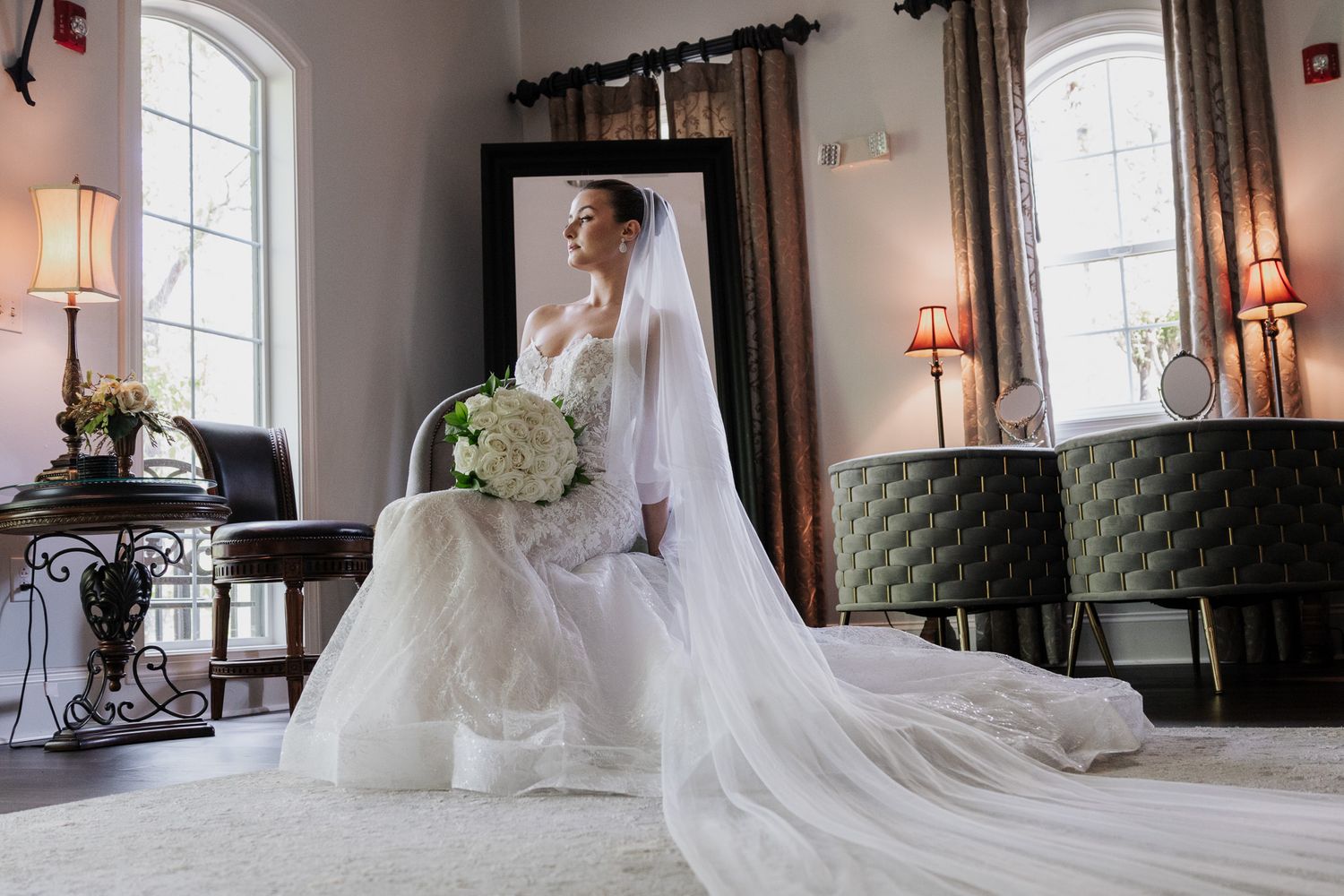 Elegant bride in a lace wedding gown with long veil holds white bouquet beside a large mirror in a bright room.
