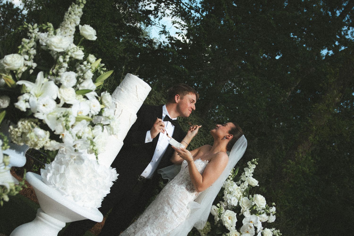 Bride and groom sharing a joyful moment beside a white tiered wedding cake adorned with white flowers outdoors.