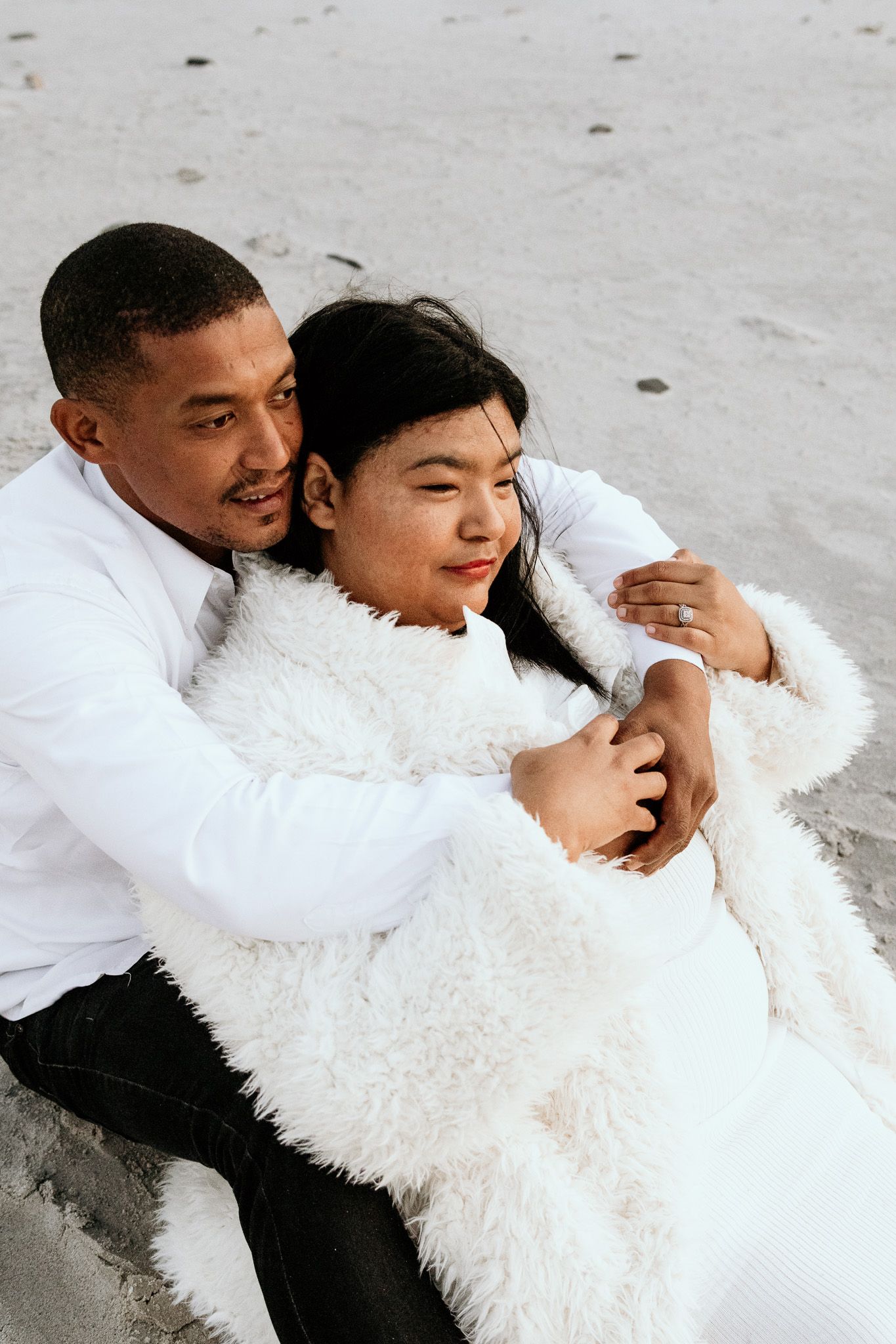 Couple sitting on beach sand embracing warmly, woman wearing diamond ring and white fur coat.