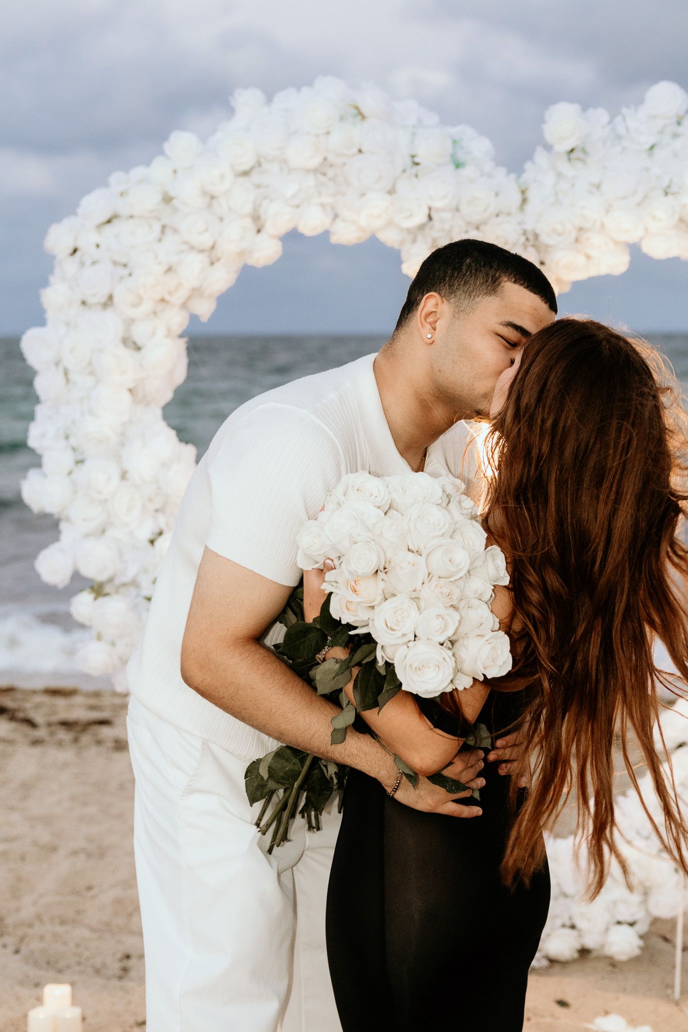 Couple kissing on beach during proposal with white floral arch and large bouquet of white roses.