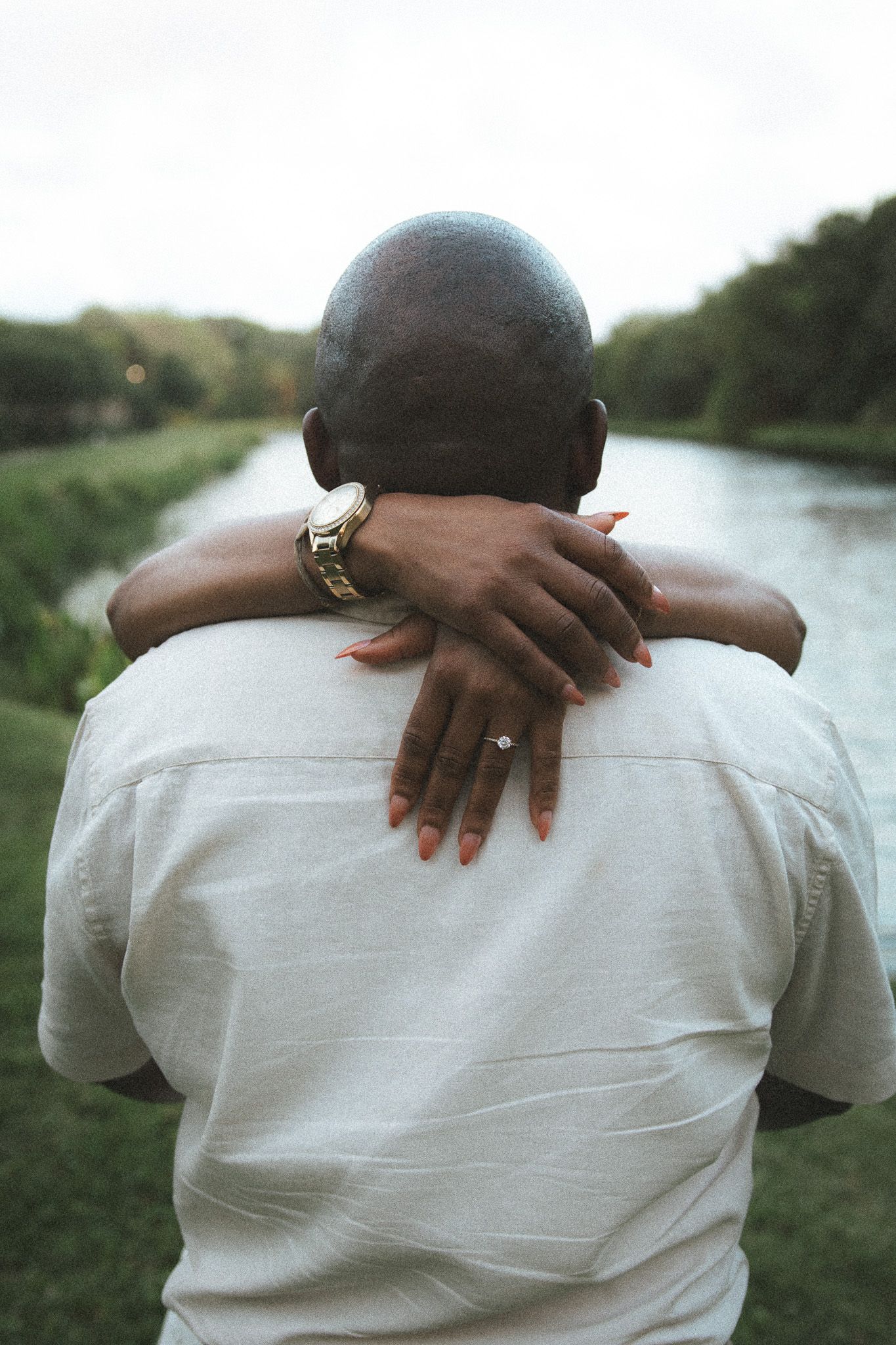 Back view of a couple embracing outdoors, woman's hands showing engagement ring and gold watch.