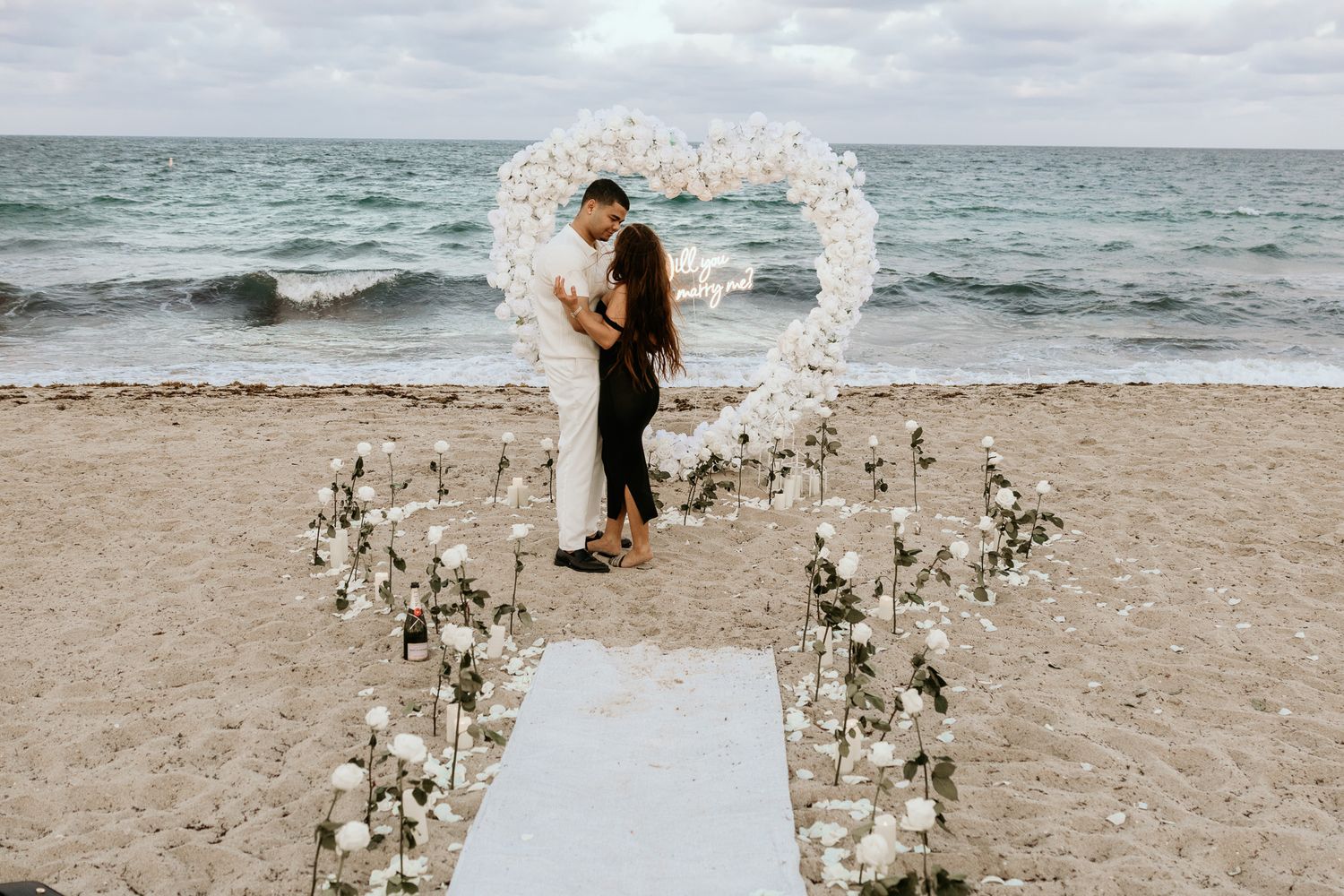 Couple embracing under a white floral heart arch on a beach with white roses lining the aisle.