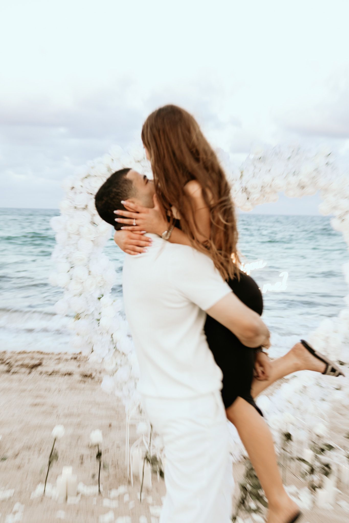 Man lifting woman for a kiss near a white floral arch on a scenic beach.