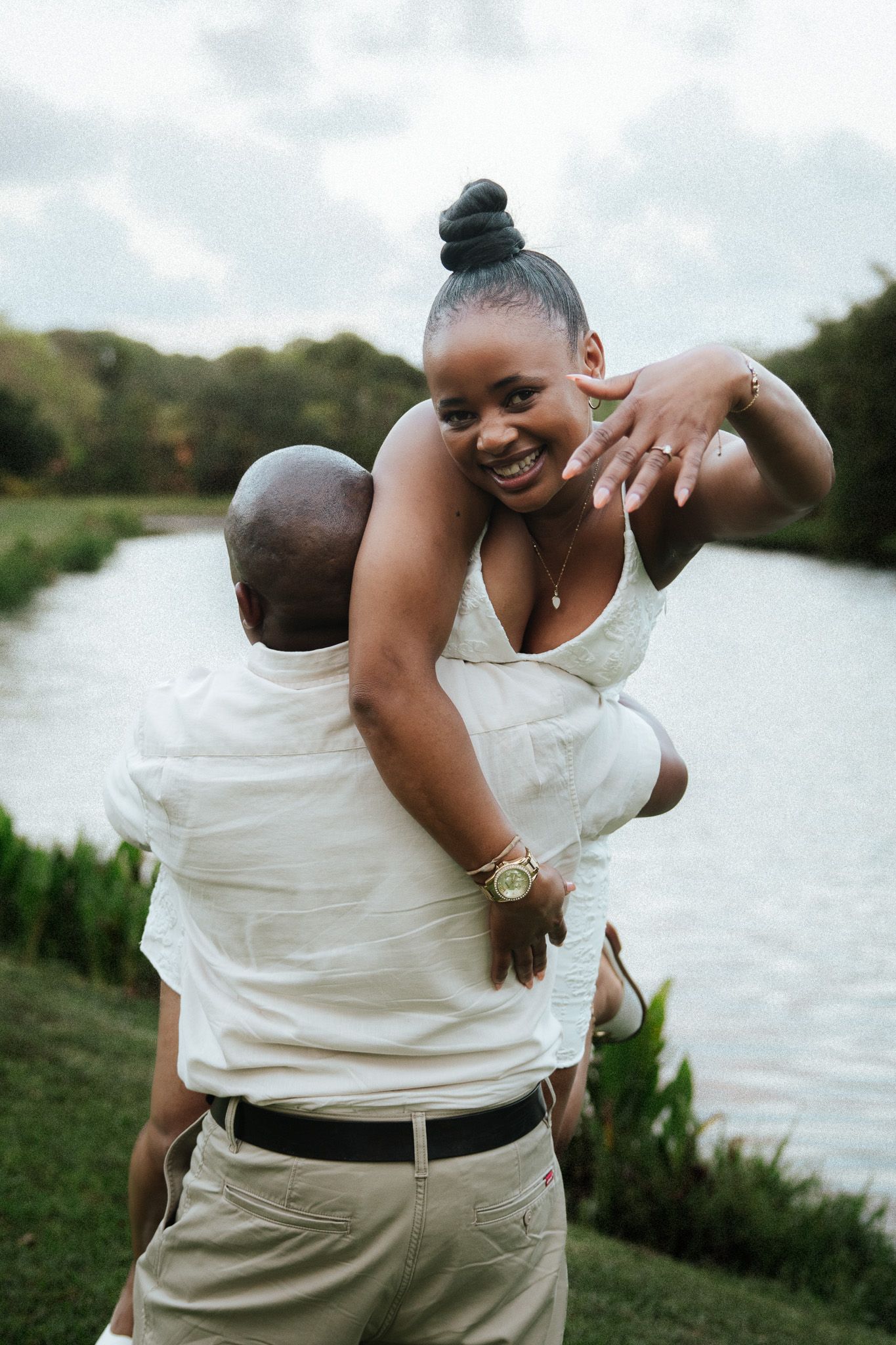 A joyful woman being lifted by her partner near a scenic pond, laughing and showing her ring.