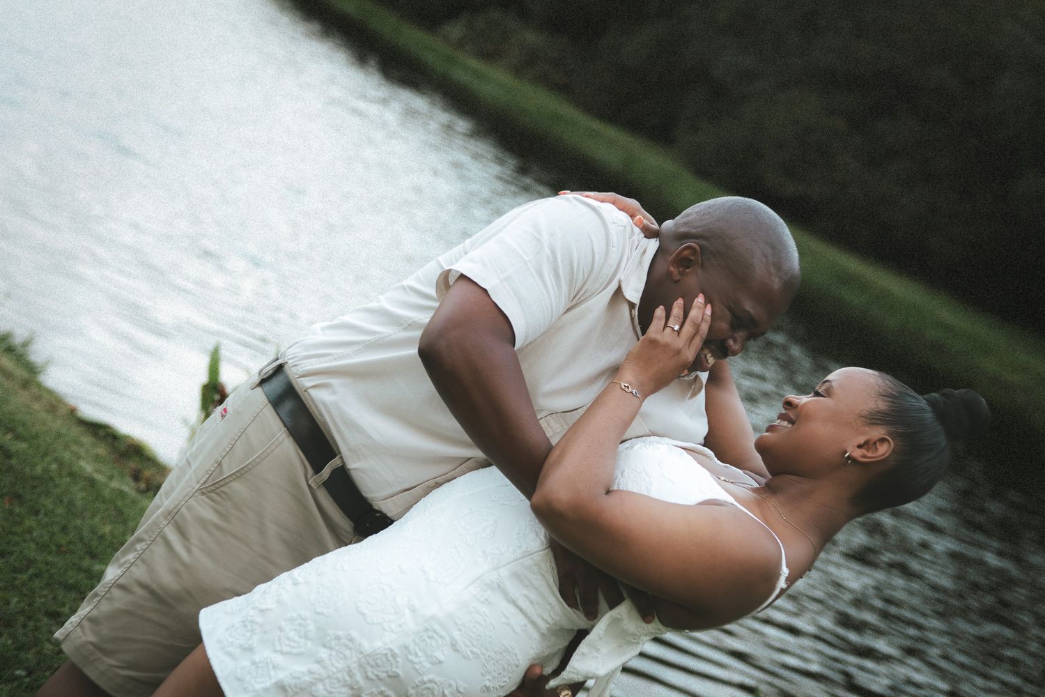 A couple sharing a romantic dip and near-kiss by a reflective pond surrounded by green trees.