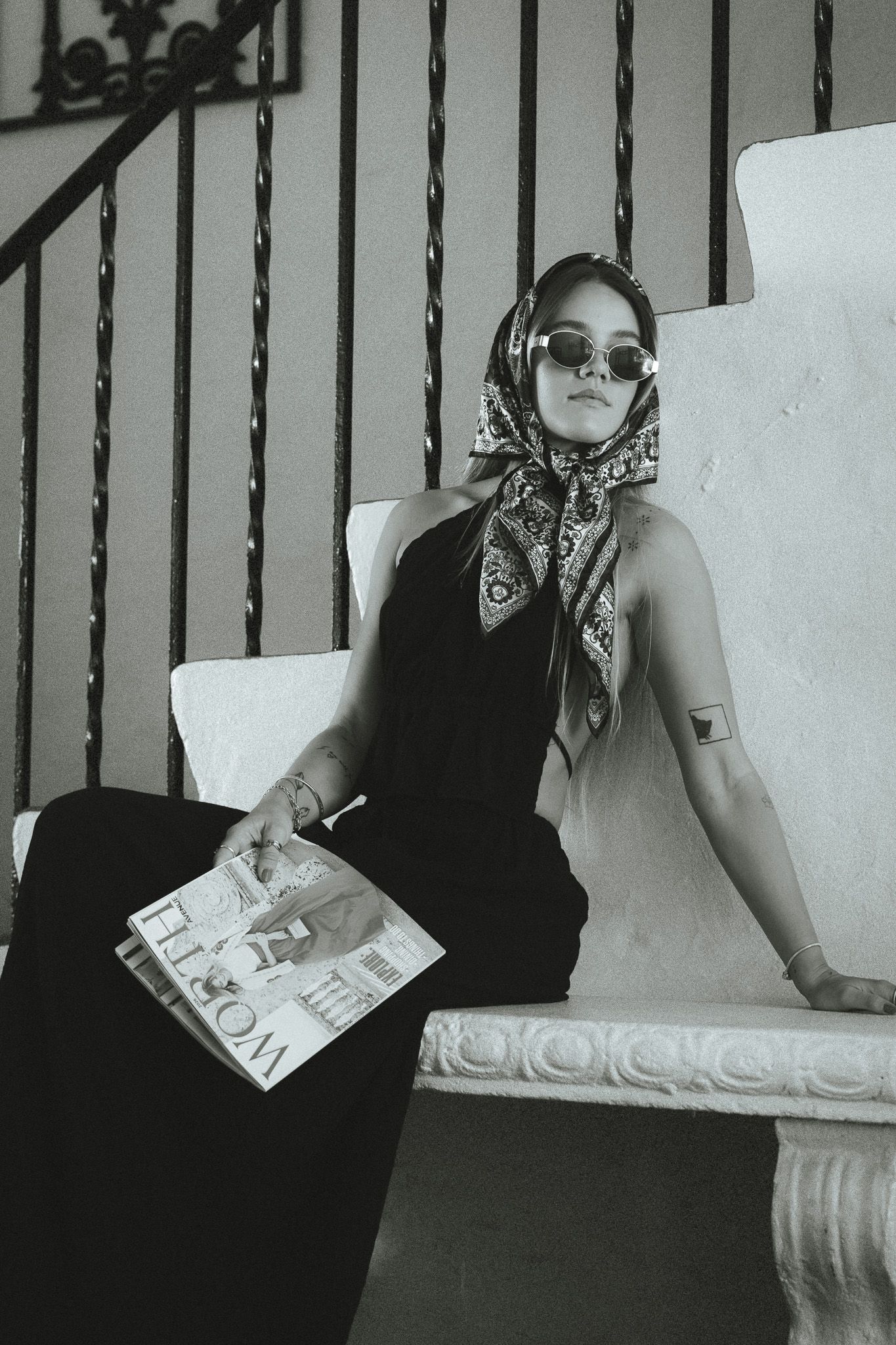 Black and white photo of a stylish woman in headscarf, sunglasses, black dress, holding a magazine while seated on a bench.