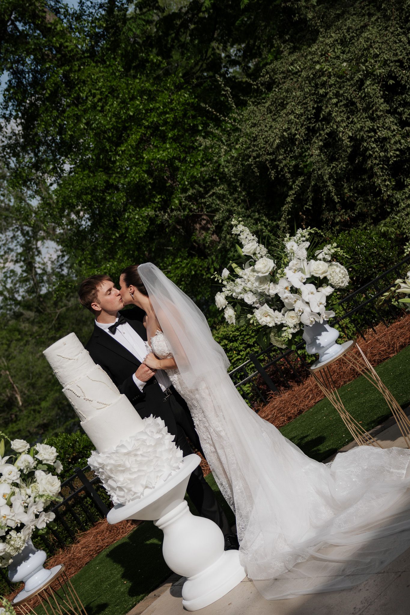 Bride and groom kiss beside a tall white layered wedding cake surrounded by elegant white floral arrangements outdoors.