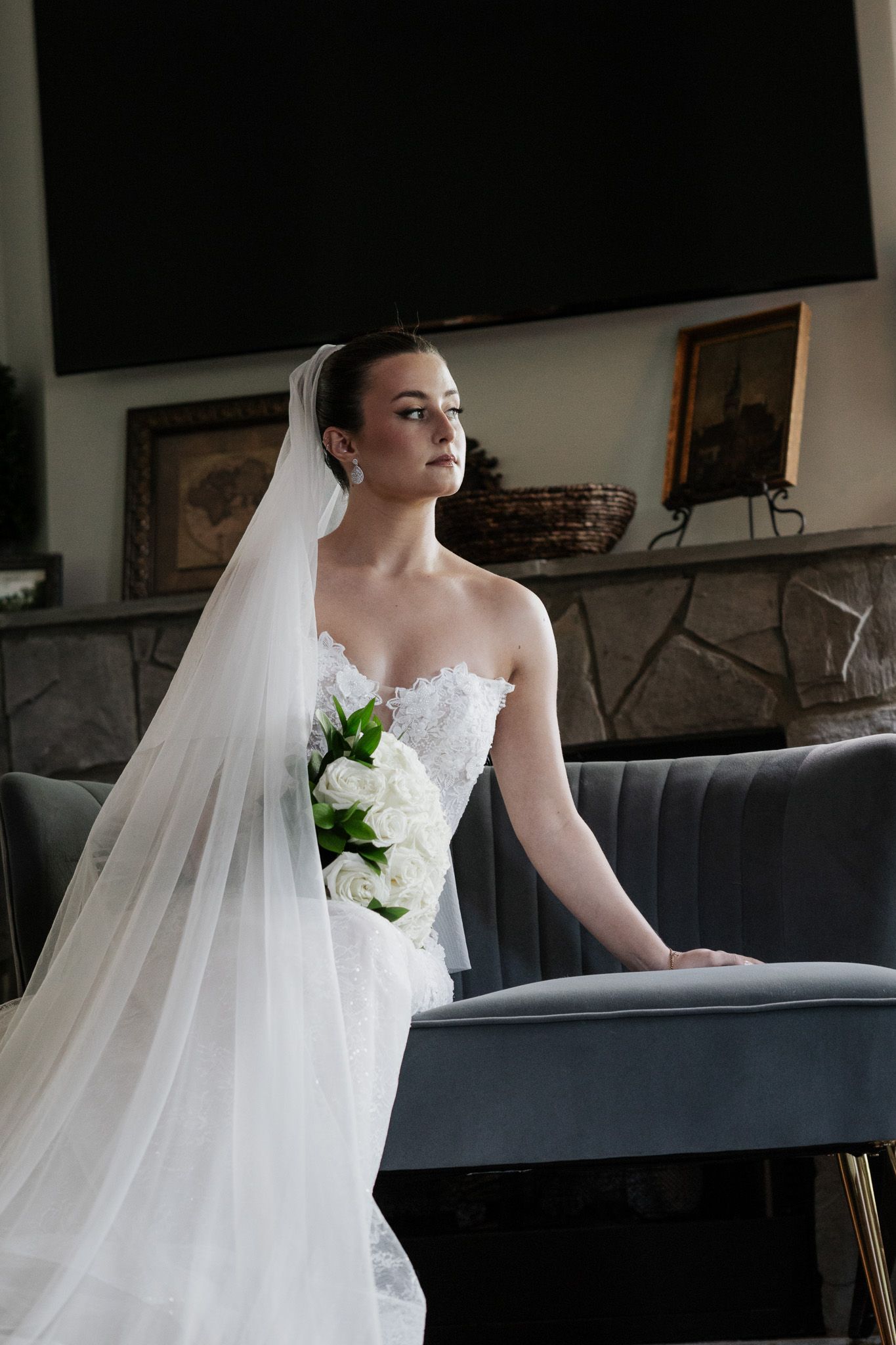 Elegant bride in a strapless lace gown with flowing veil holds white bouquet while seated near a stone fireplace.