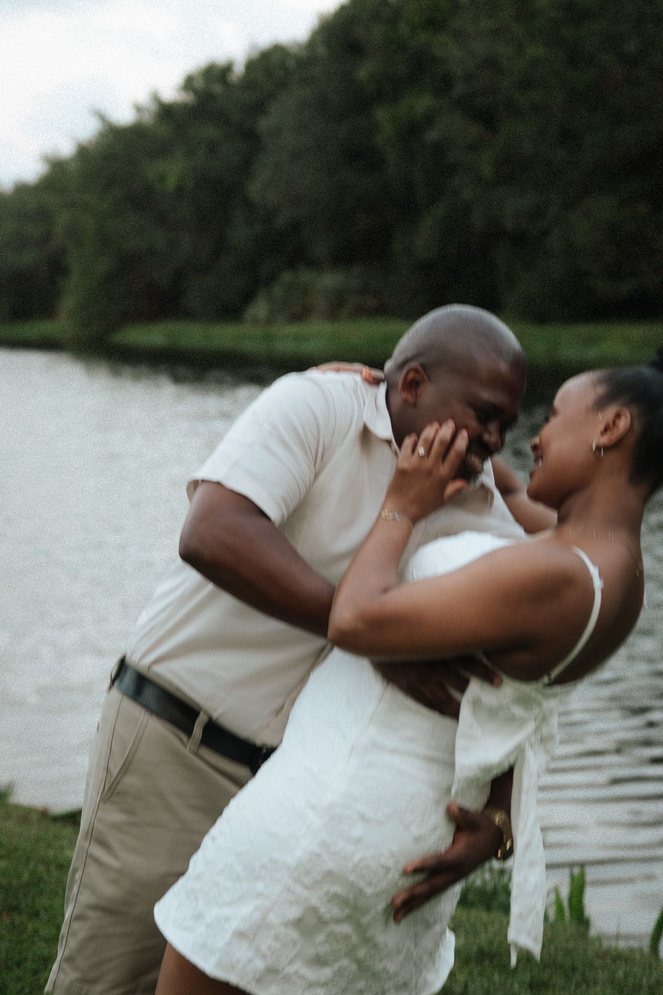 A couple shares a tender moment by a lake, the man kissing the woman's hand as she smiles in a white dress.