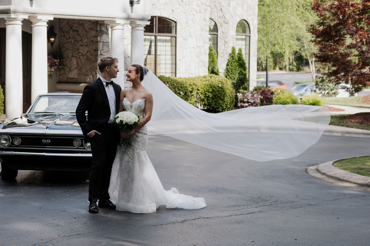 Bride and groom pose beside a classic black car, her veil flowing in the wind outside an elegant venue.