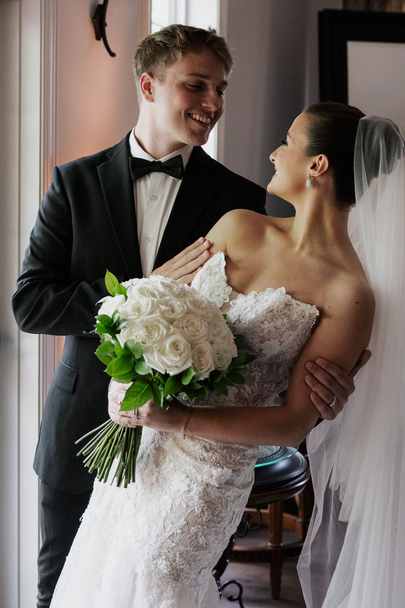 Smiling bride and groom sharing joyful moment indoors, bride holding white rose bouquet in lace wedding dress.