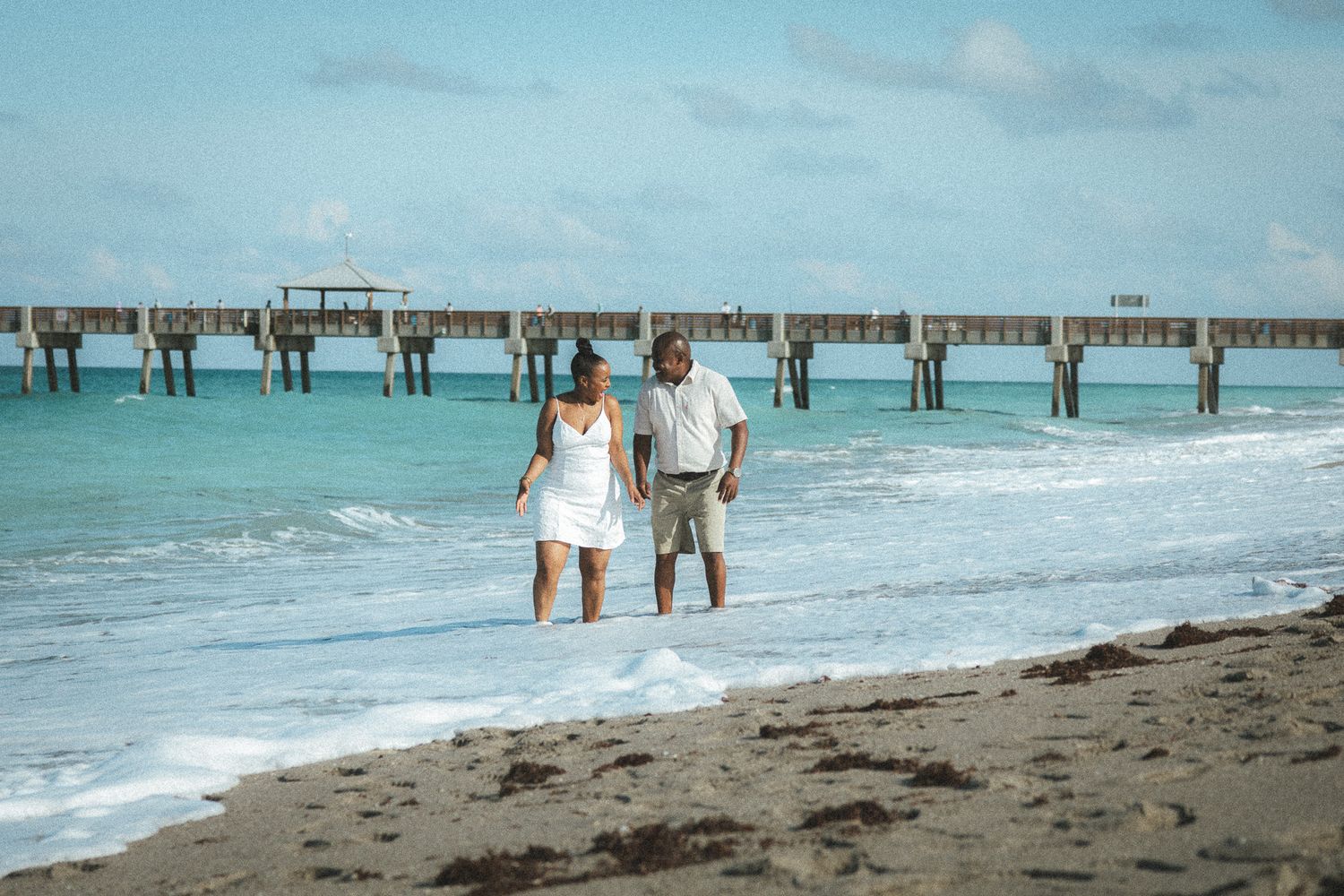 Couple in white outfits walk barefoot along a tropical beach shoreline with a wooden pier stretching into the ocean.