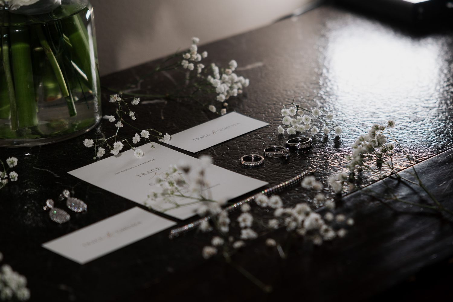 Elegant wedding stationery with rings, earrings, and baby's breath flowers on a dark table.
