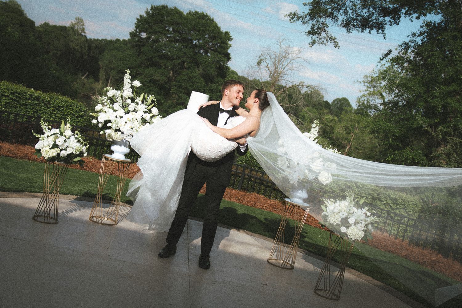 Groom carries bride in flowing white veil at outdoor wedding surrounded by lush green landscape.