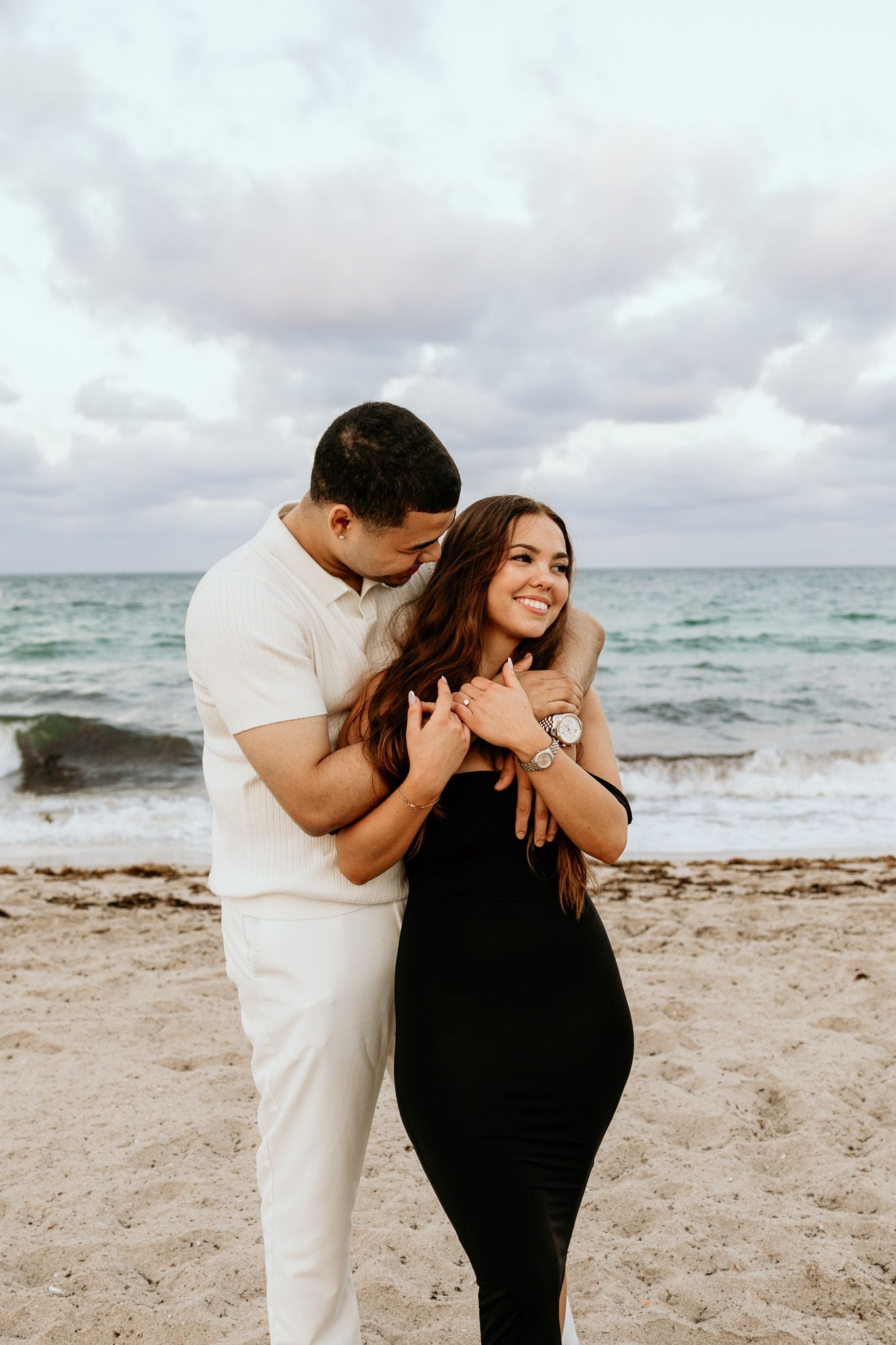 Couple holding hands and smiling on a sandy beach with cloudy ocean backdrop.