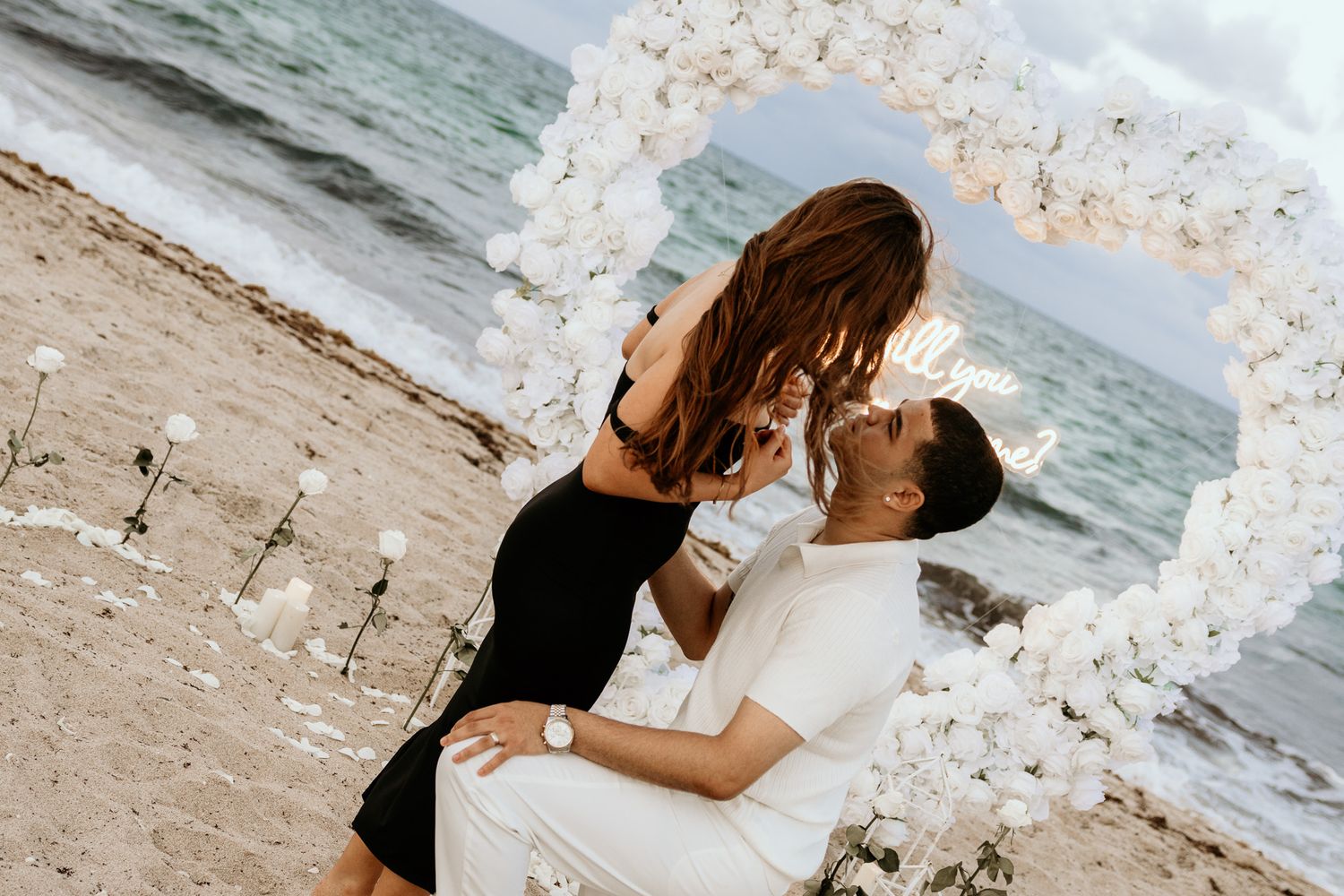 Couple sharing a romantic kiss under a white floral arch on a sandy beach.