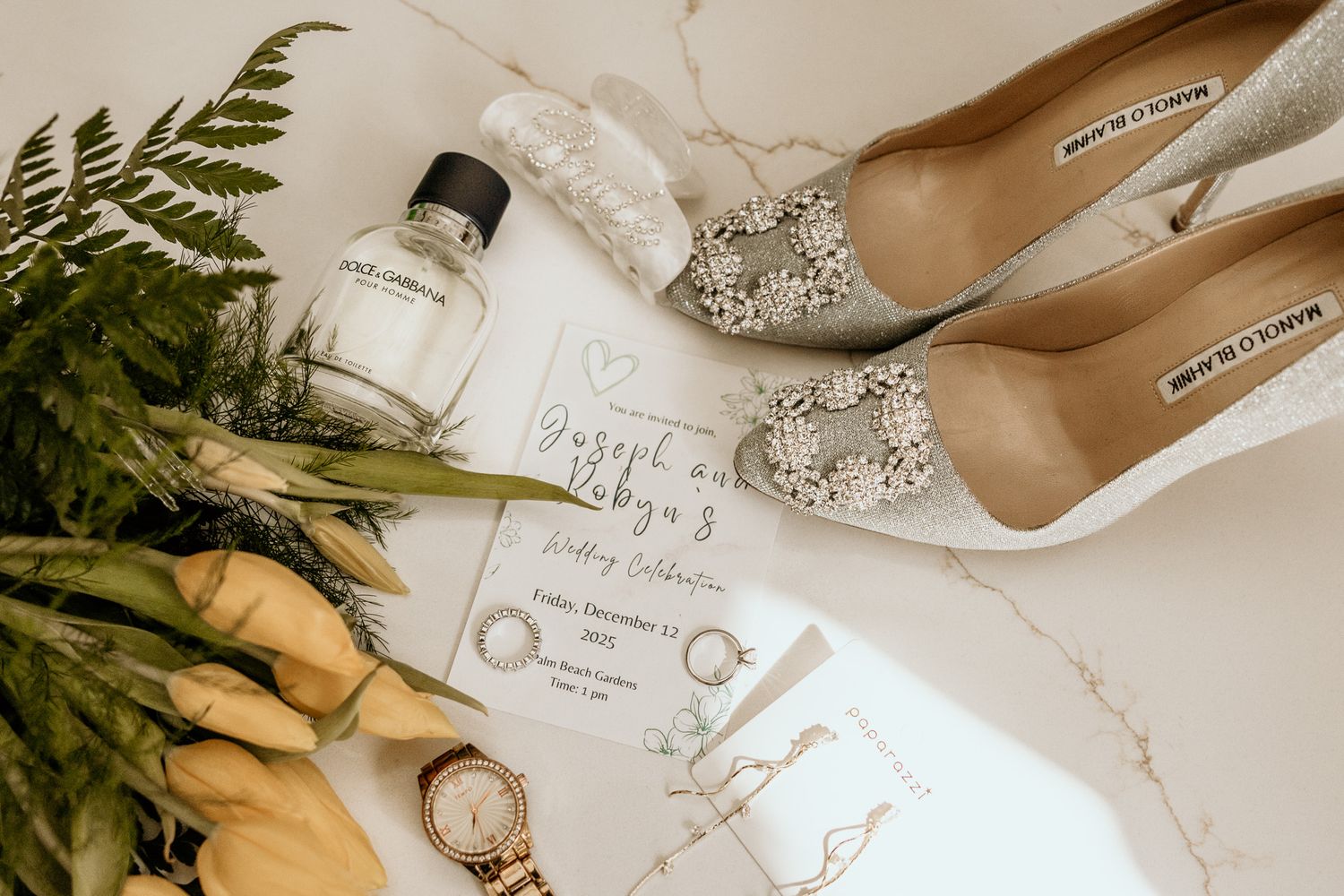 Bridal flat lay featuring silver embellished heels, yellow tulips, perfume, watch, and wedding invitation.