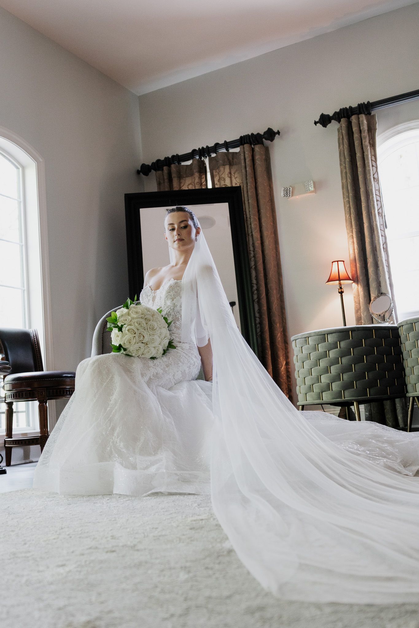 Full-length bridal portrait in lace gown with cathedral veil trailing across white carpet near mirror.