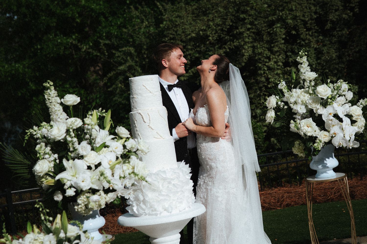 Bride and groom smile together beside a tall white wedding cake with lush floral arrangements.