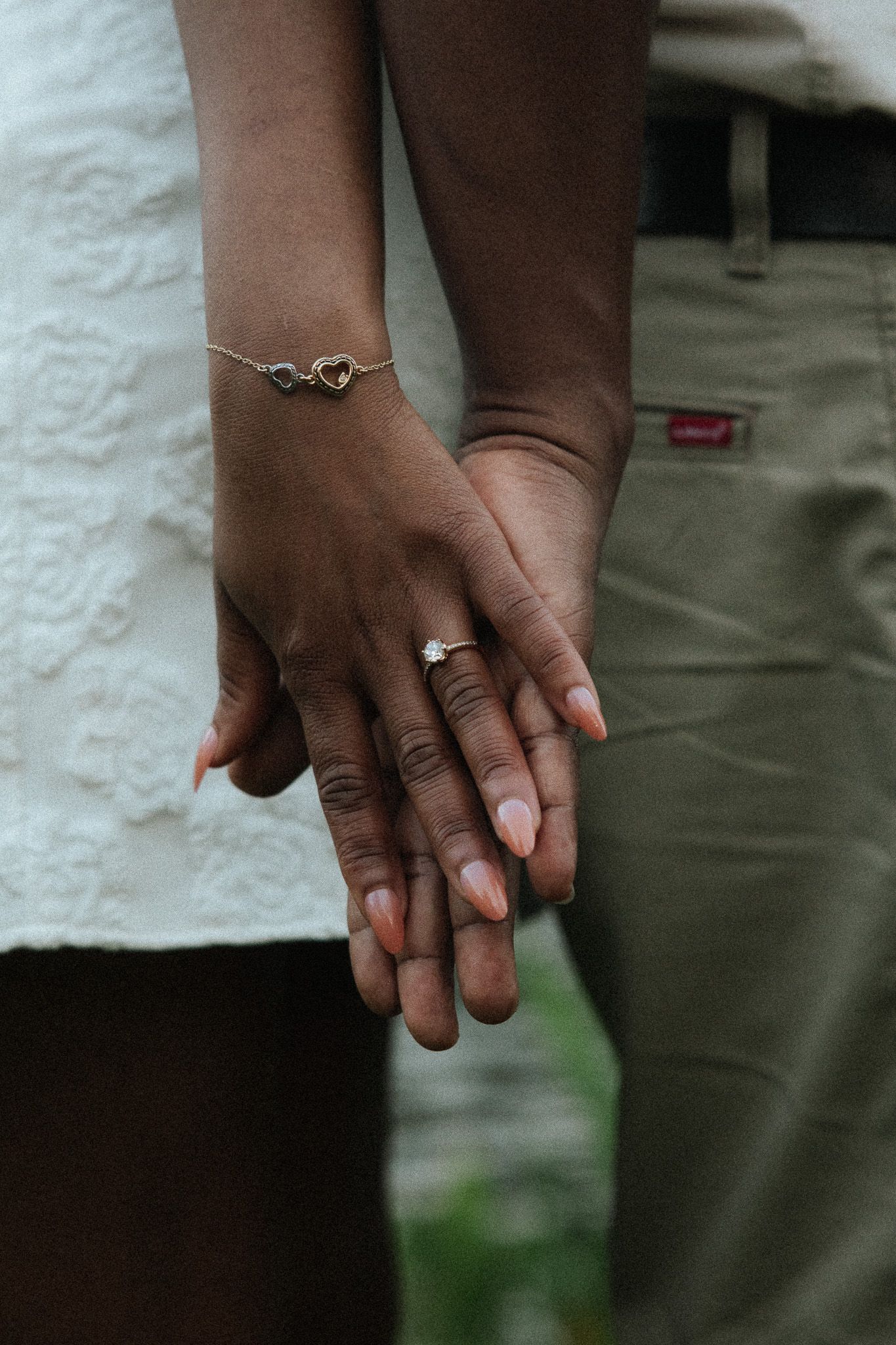 A close-up of an engaged couple holding hands, displaying a diamond ring and delicate heart bracelet.
