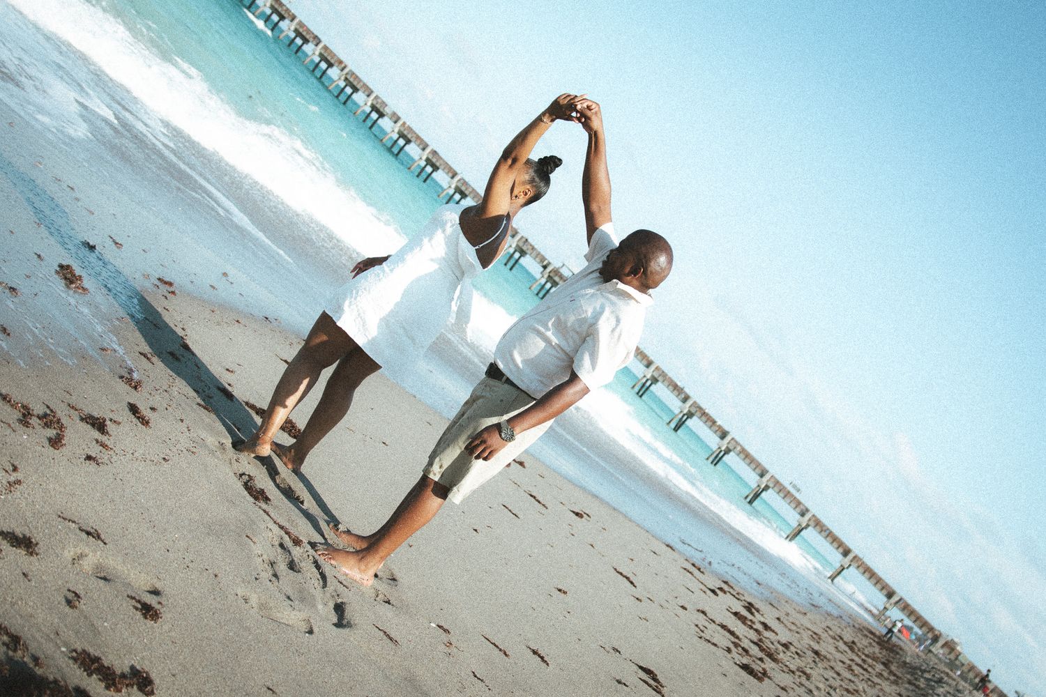 Couple in white outfits pose playfully on a sunny beach, leaning away from each other while holding hands by the ocean.