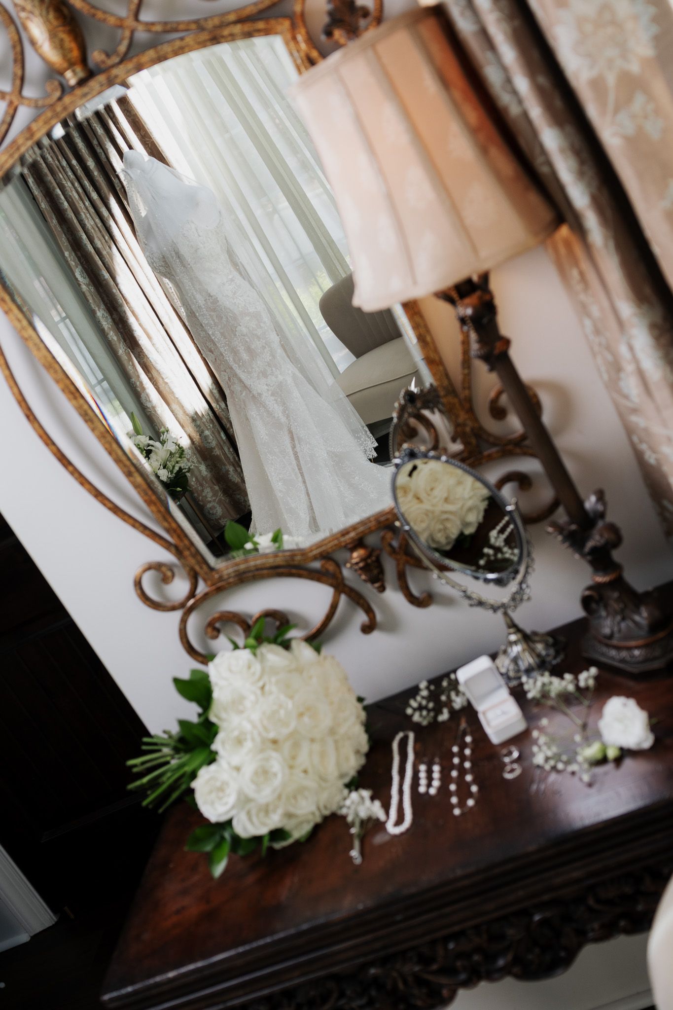 Bridal preparation scene with wedding dress reflected in ornate mirror beside white rose bouquet and jewelry.