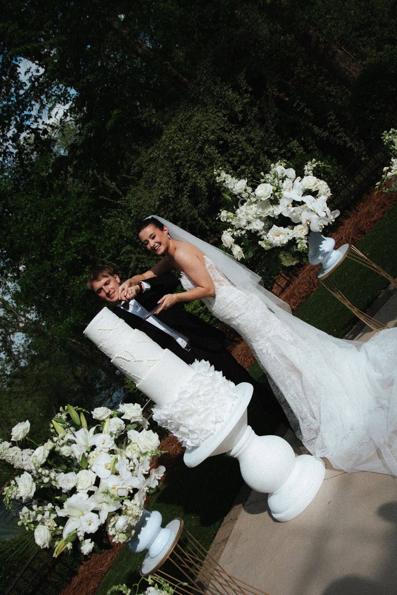 Aerial view of bride and groom cutting elegant white tiered wedding cake surrounded by white floral arrangements.