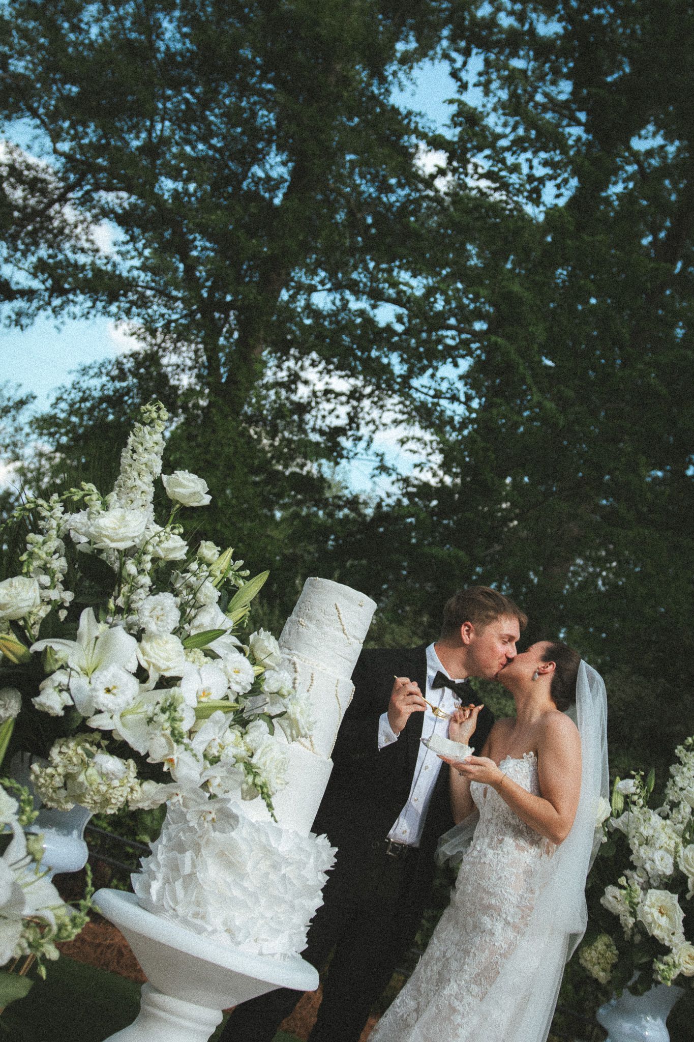 Newlyweds kissing while holding cake slices beside a white tiered wedding cake with lush white floral arrangements.