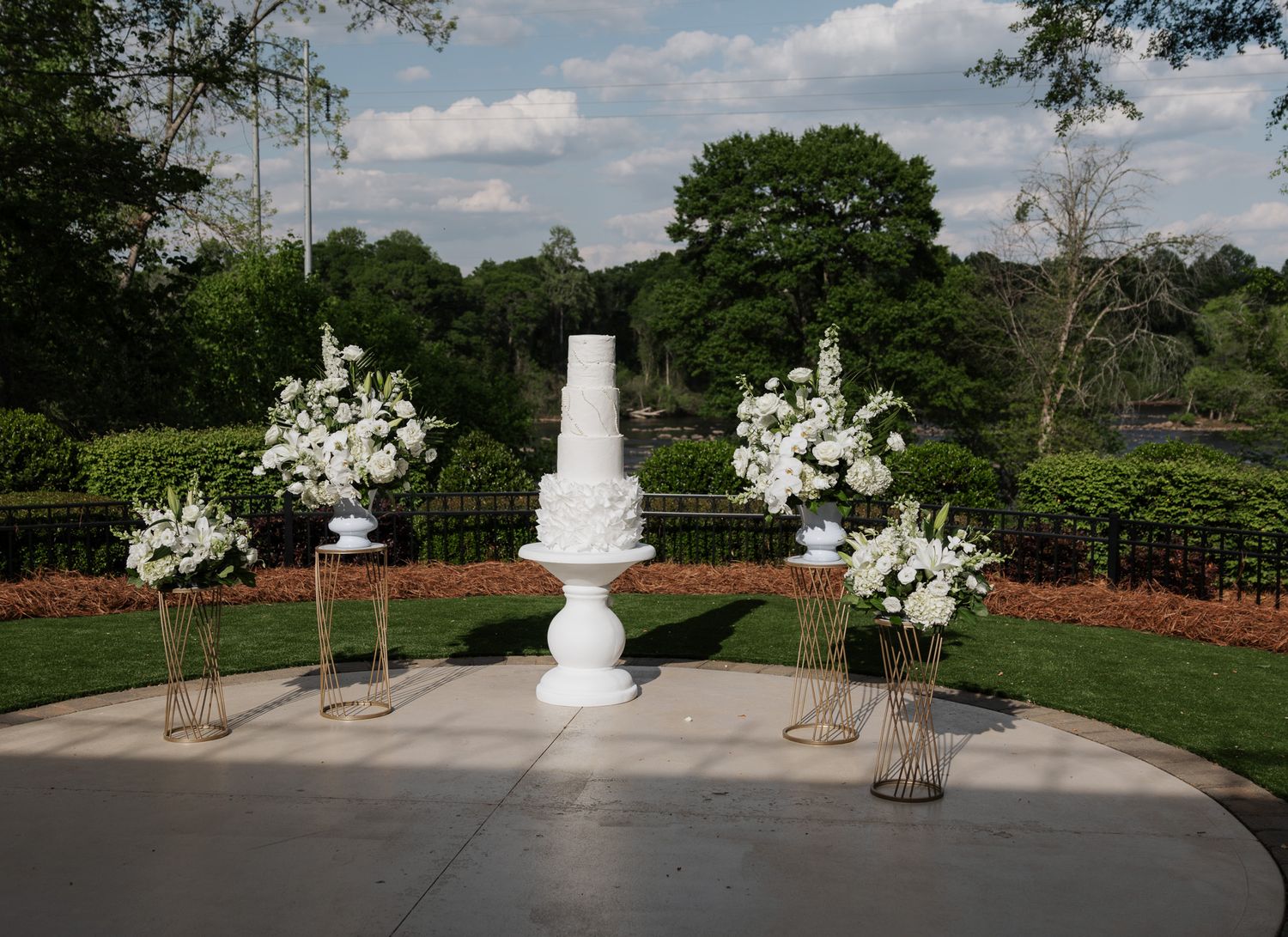 Elegant white tiered wedding cake displayed outdoors surrounded by white floral arrangements.