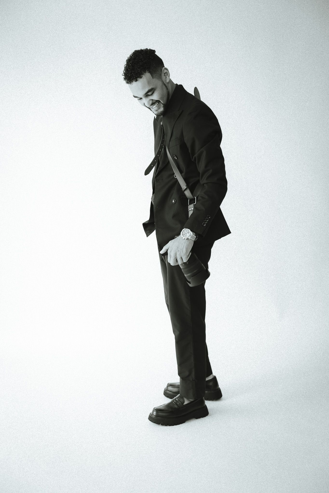 Black and white photo of a stylish man in a dark suit and chunky loafers, posing with a slight lean and gloved hand.