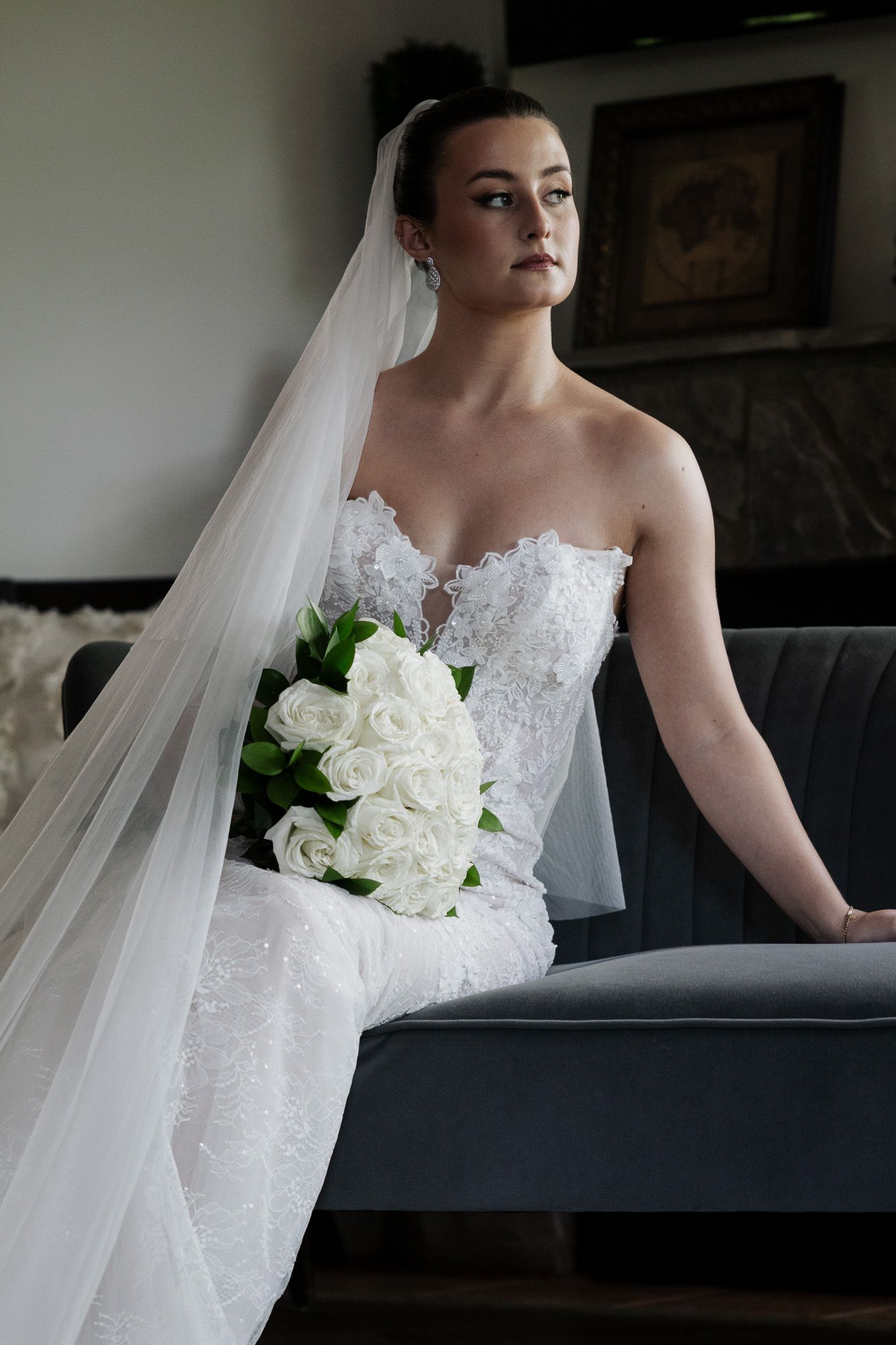 Stunning bride in strapless lace wedding dress with cathedral veil holds white roses while seated on a gray couch.
