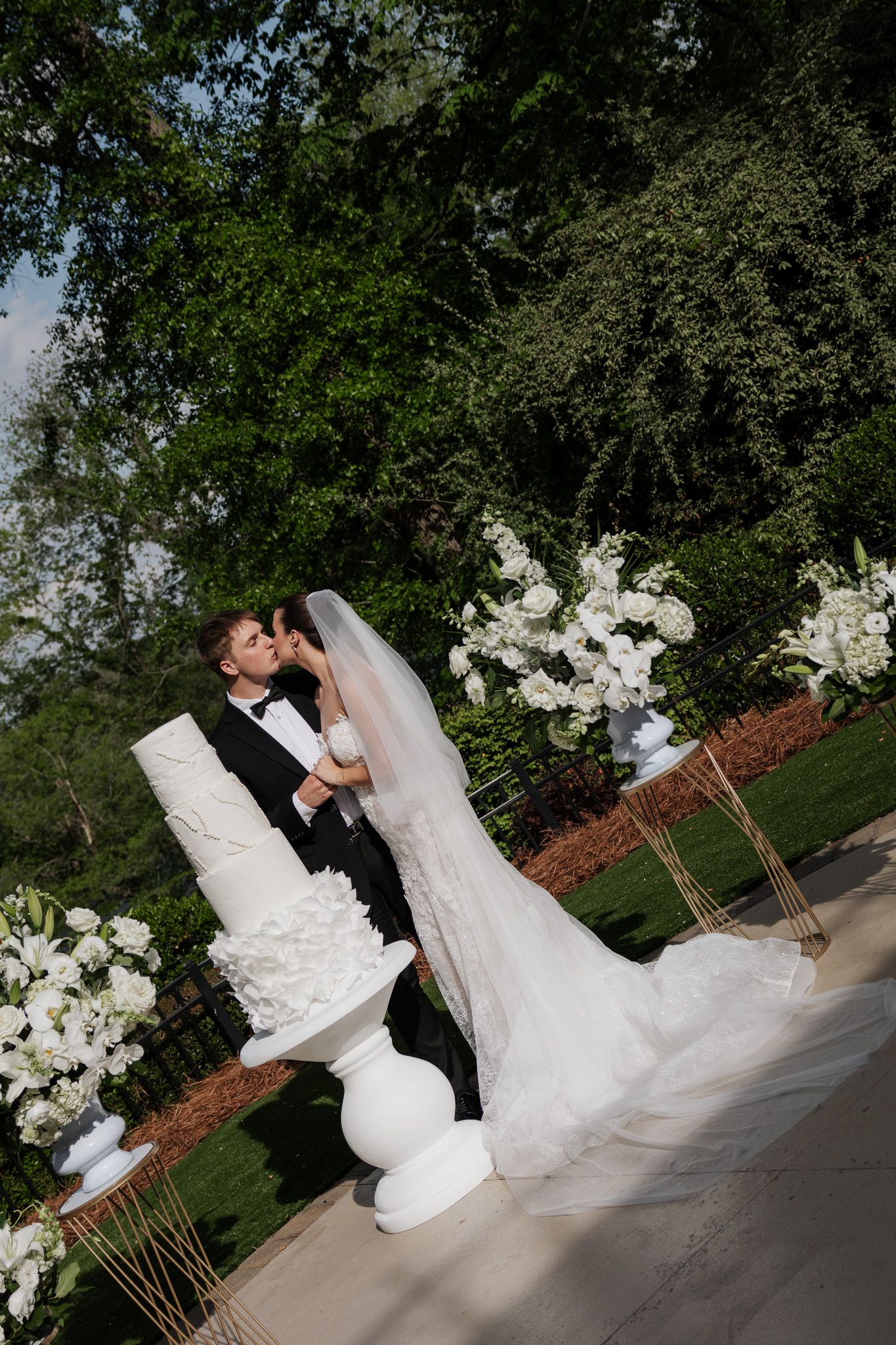 Newlyweds share a passionate kiss next to their tall white textured wedding cake amid lush garden floral decor.