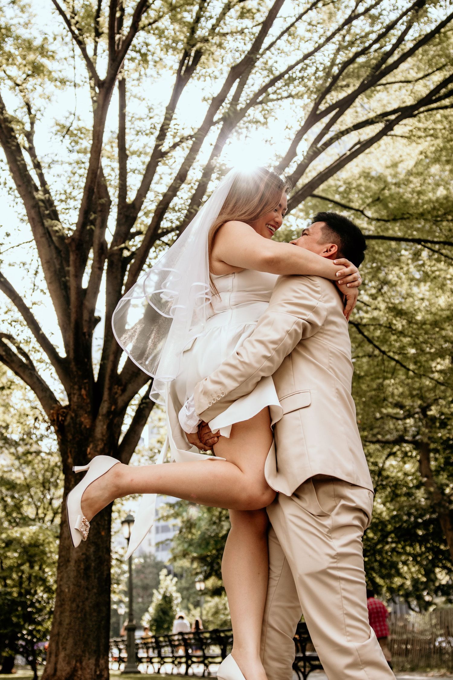Groom lifts smiling bride in white mini dress and veil in a sunlit park with tall trees.