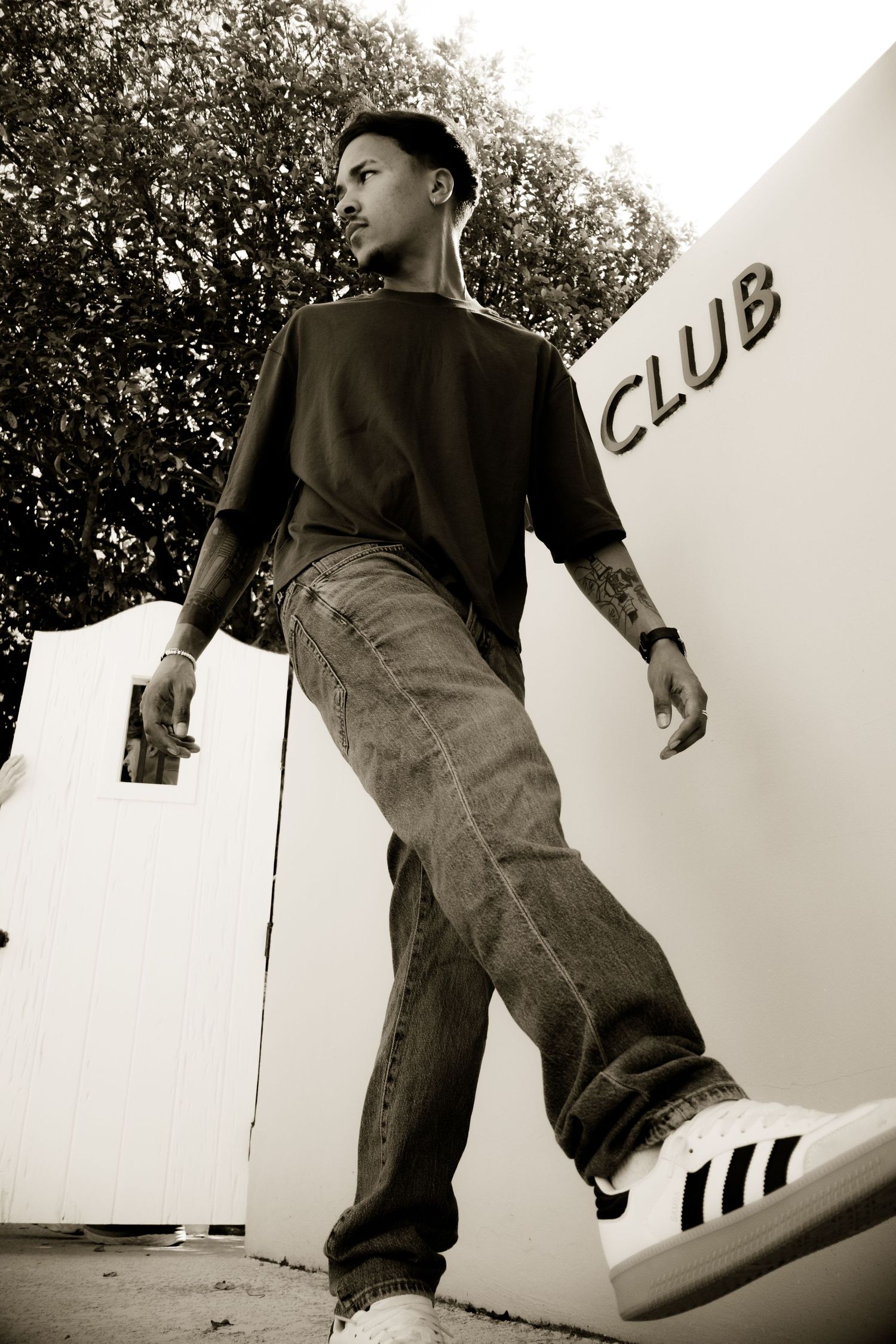 Young tattooed man in baggy jeans and Adidas sneakers posing near a club entrance.
