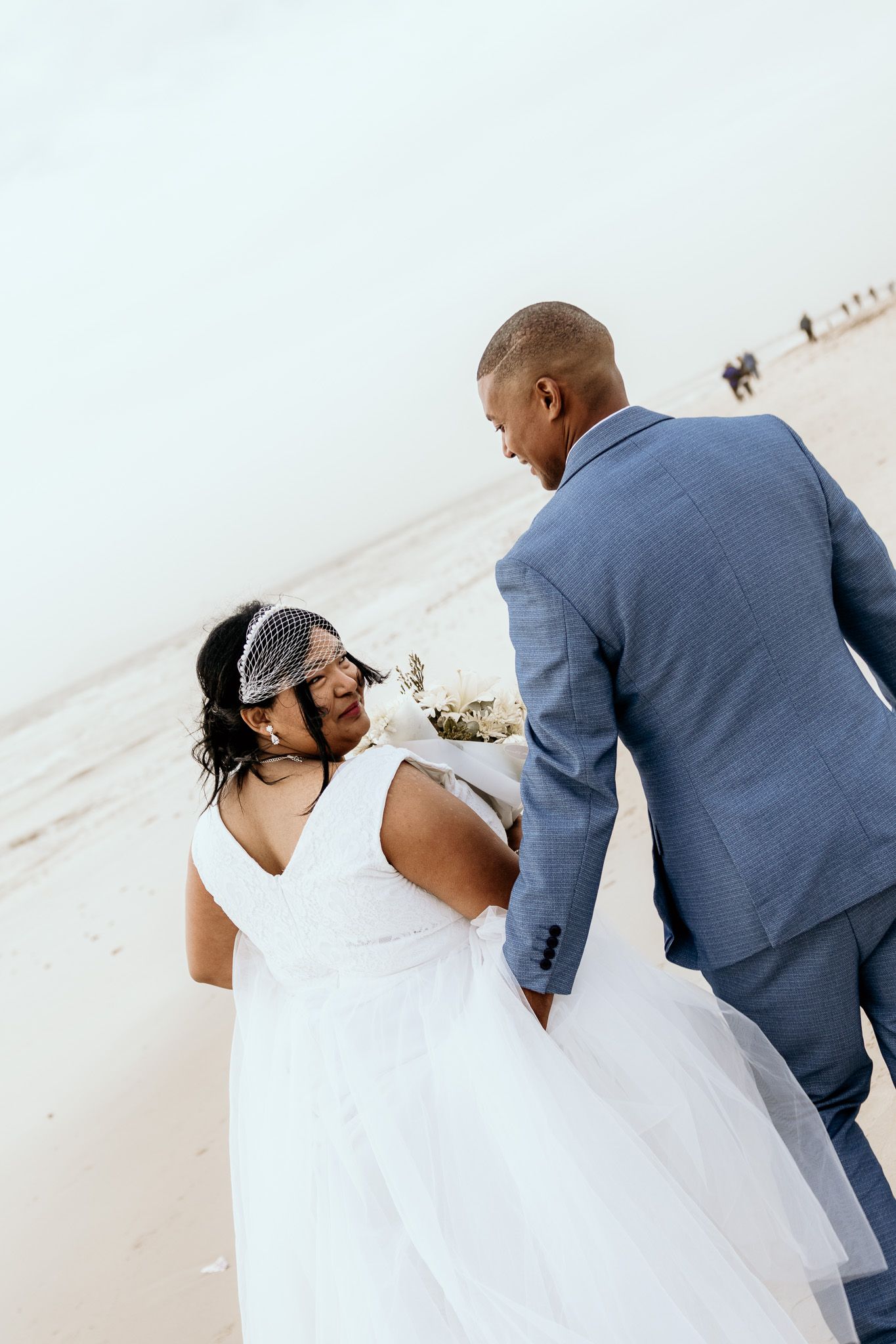 Bride in white gown and birdcage veil walks hand in hand with groom in blue suit on beach.
