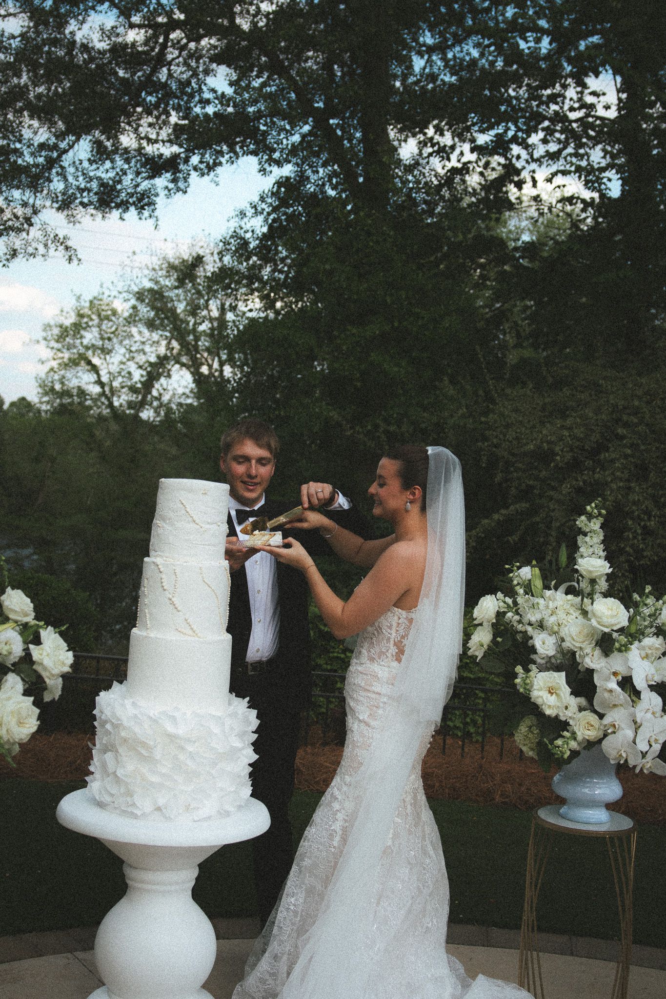 Bride and groom cutting a tall white four-tier wedding cake outdoors surrounded by white floral arrangements.
