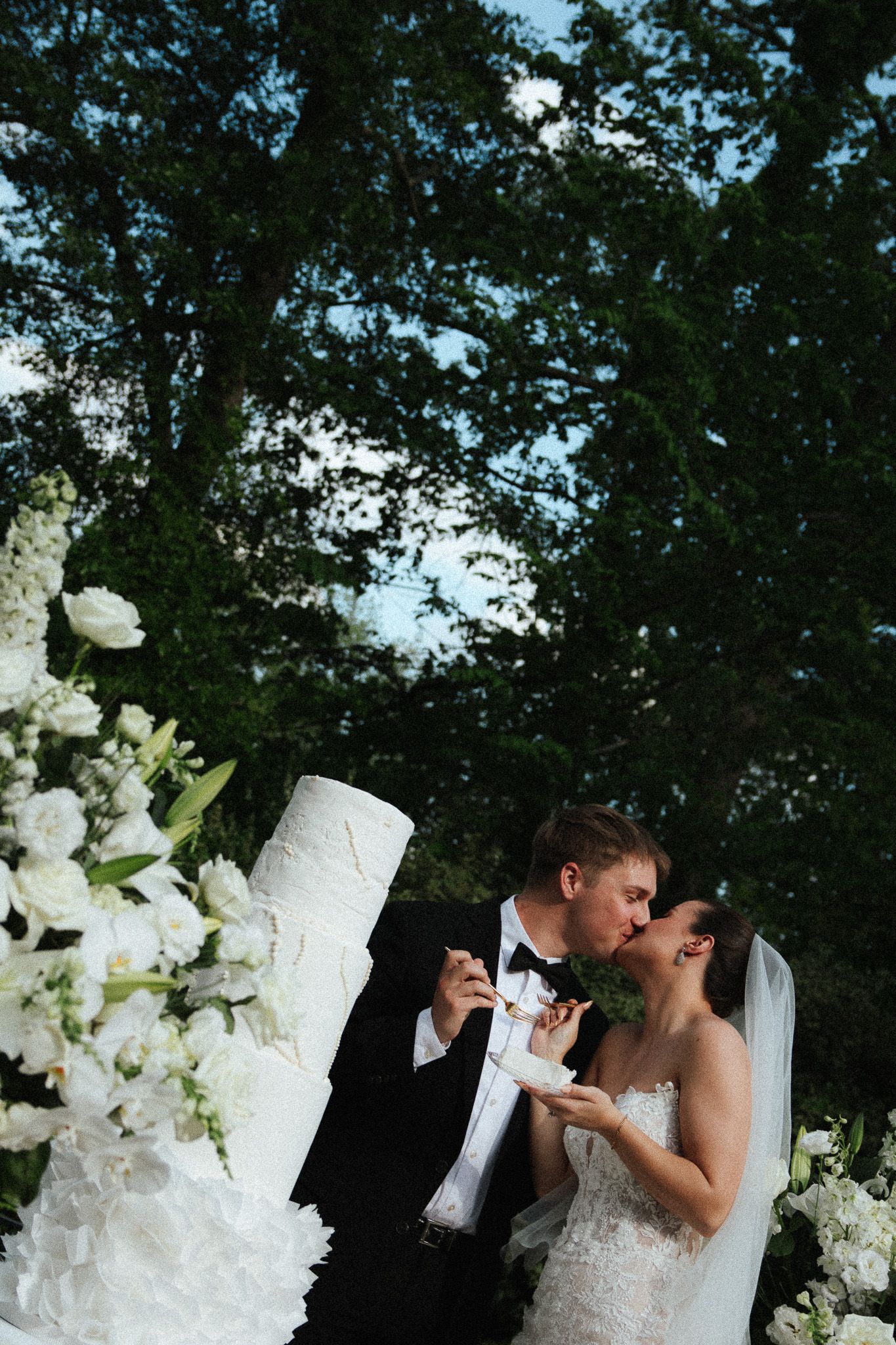 Newlyweds share a romantic kiss while holding wedding cake slices beside a white tiered cake outdoors.