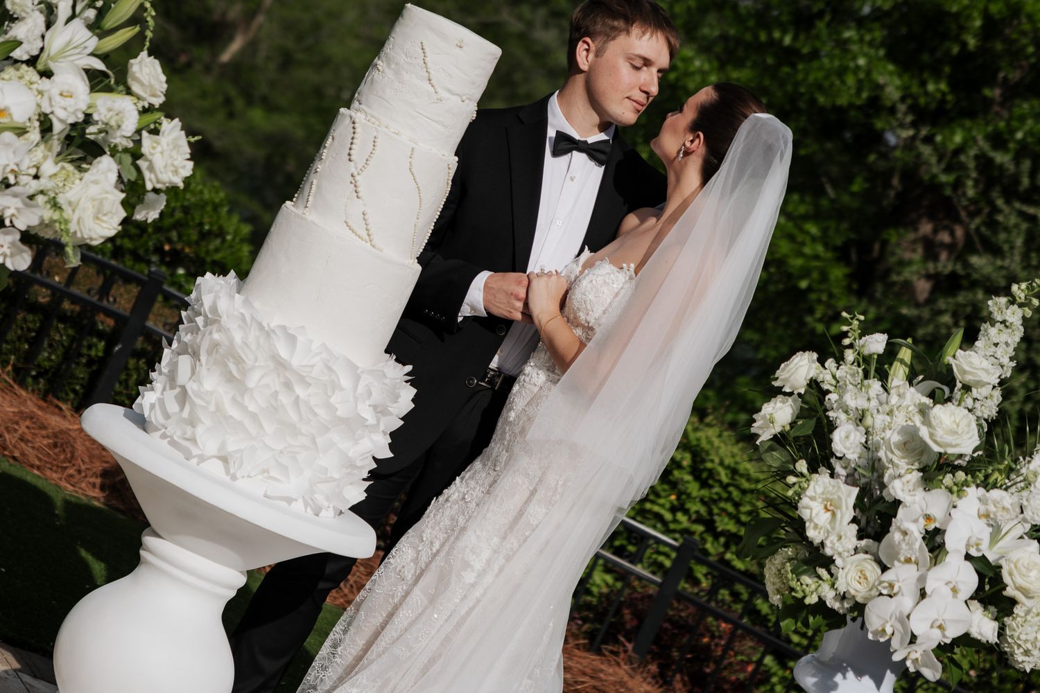 Bride and groom share a kiss while cutting their elegant white multi-tiered wedding cake outdoors among white florals.
