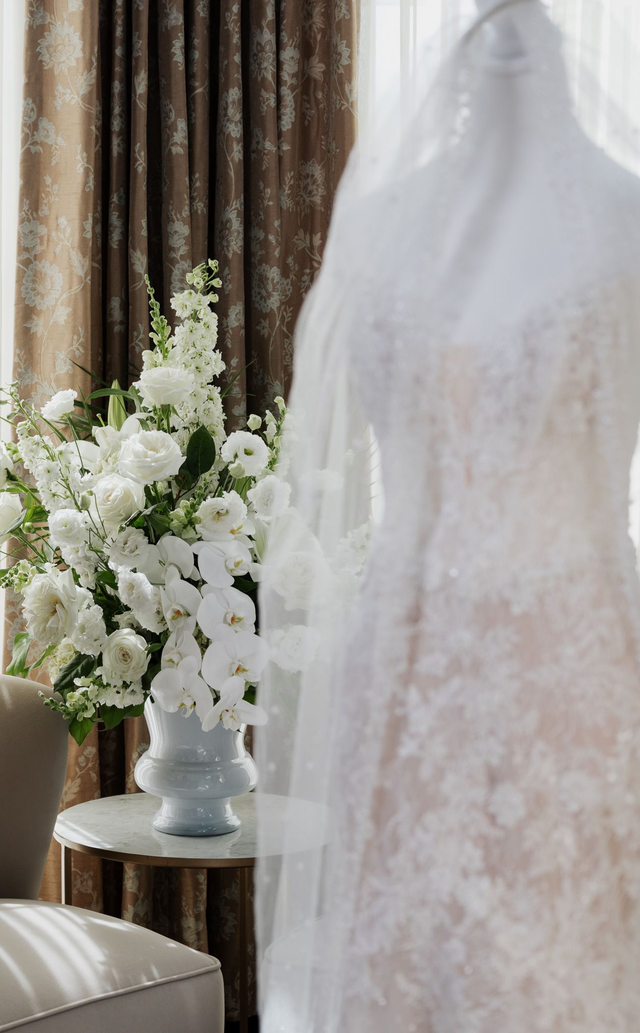 Lace wedding dress on mannequin beside white floral arrangement in a luxurious hotel room.